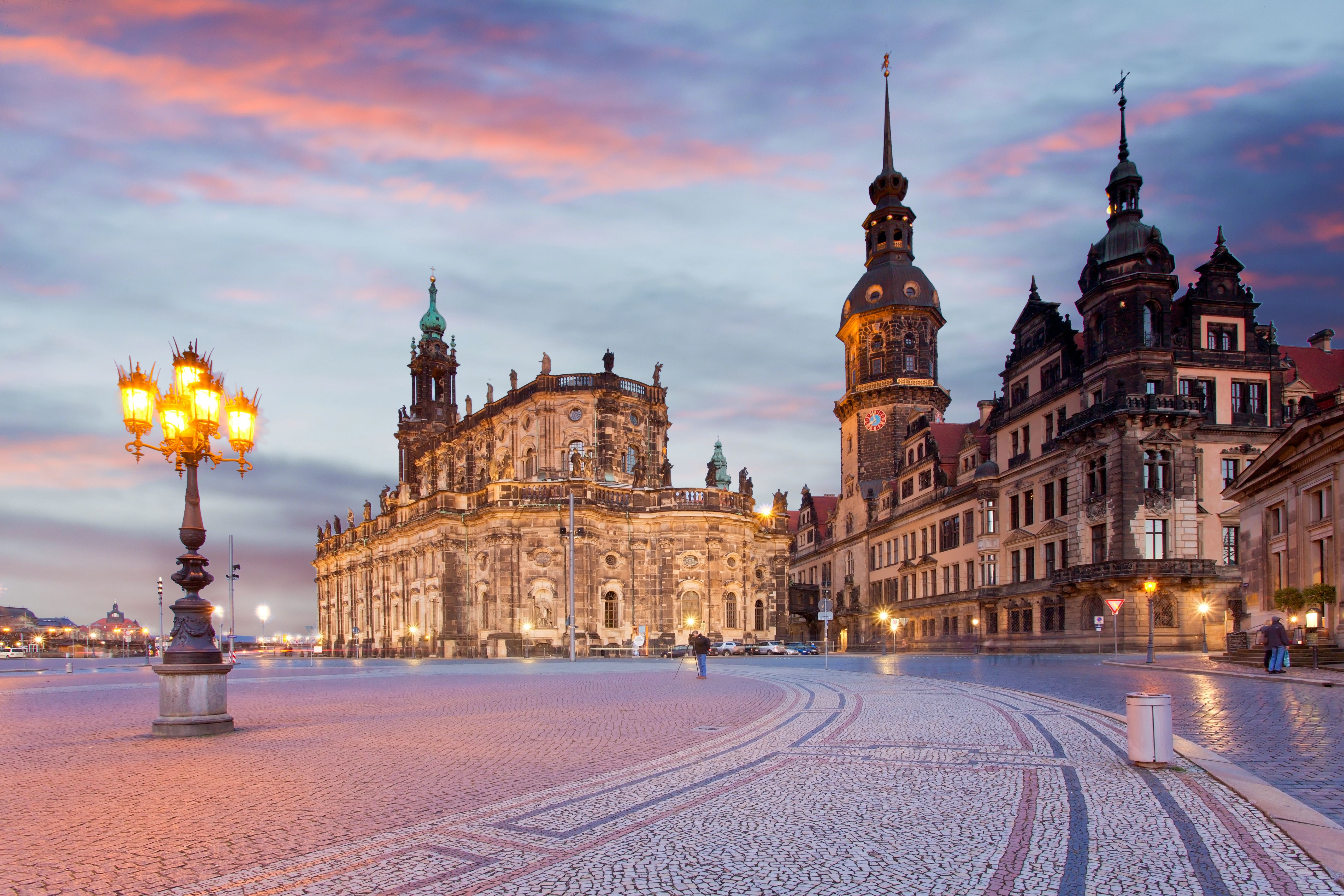 Abendlicher Blick auf das historische barocke Gebäude der Katholischen Hofkirche und angrenzende Bauten in Dresden, mit beleuchteten Laternen und einem farbenfrohen Himmel im Hintergrund.