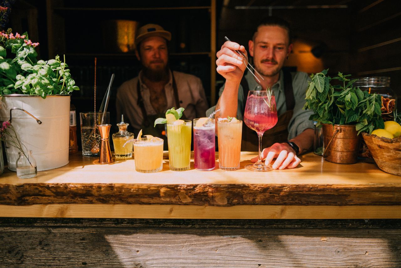 Zwei Barkeeper bereiten an einer Bar fünf farbenfrohe Cocktails zu, umgeben von frischen Kräutern und stilvoller Dekoration. Zwei Barkeeper bereiten an einer Bar fünf farbenfrohe Cocktails zu, umgeben von frischen Kräutern und stilvoller Dekoration.