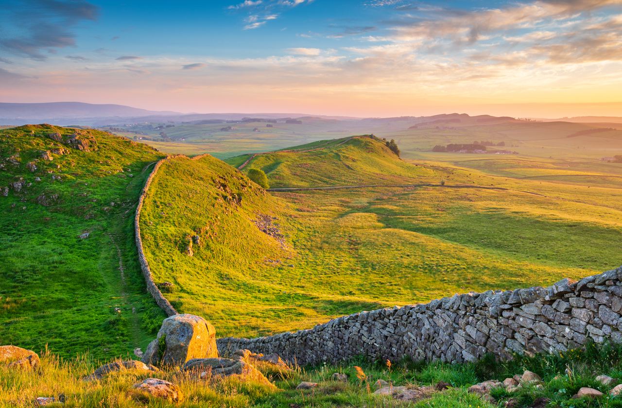 Weite grüne Hügel im Abendlicht, durchzogen von einer historischen, aus Natursteinen gefertigten Mauer, die sich entlang der Landschaft schlängelt. Ruhige, ländliche Atmosphäre mit weitem Blick in den sanft gebogenen Talraum unter einem farbenfrohen Himmel. Weite grüne Hügel im Abendlicht, durchzogen von einer historischen, aus Natursteinen gefertigten Mauer, die sich entlang der Landschaft schlängelt. Ruhige, ländliche Atmosphäre mit weitem Blick in den sanft gebogenen Talraum unter einem farbenfrohen Himmel.