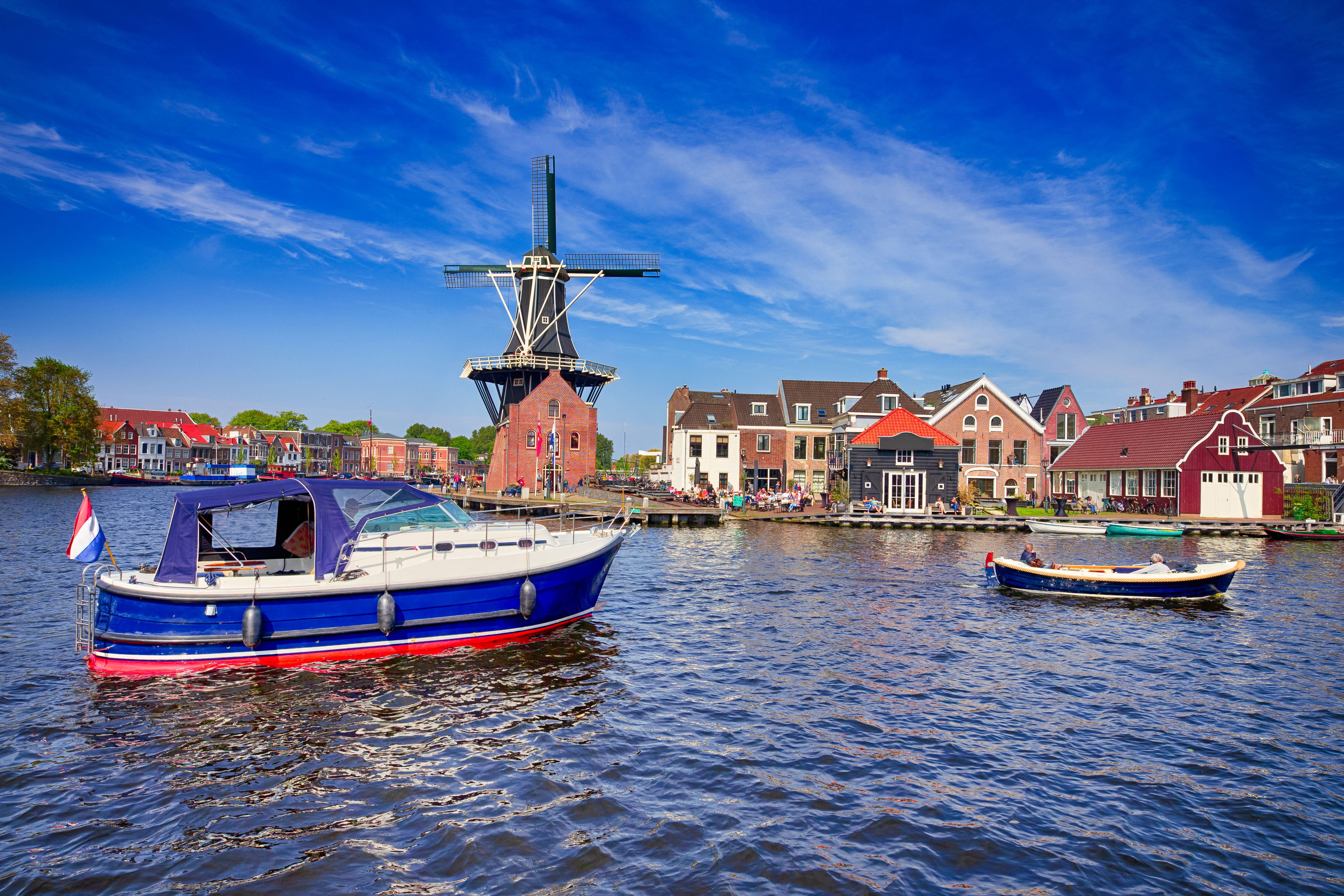 Traditionelle holländische Windmühle am Ufer eines ruhigen Kanals, umgeben von historischen Häusern und zwei Booten auf dem Wasser, unter einem klaren blauen Himmel.