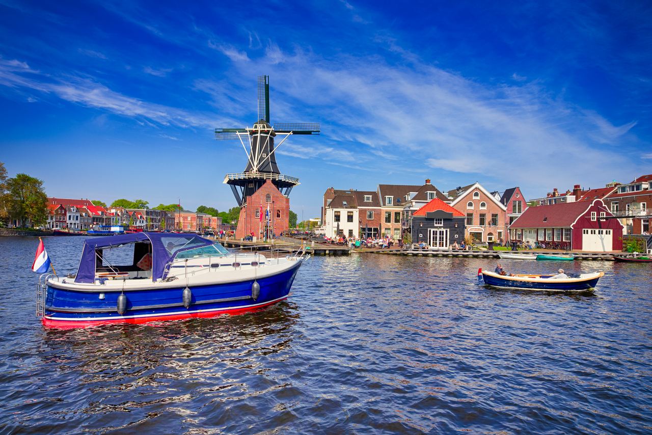Traditionelle holländische Windmühle am Ufer eines ruhigen Kanals, umgeben von historischen Häusern und zwei Booten auf dem Wasser, unter einem klaren blauen Himmel. Traditionelle holländische Windmühle am Ufer eines ruhigen Kanals, umgeben von historischen Häusern und zwei Booten auf dem Wasser, unter einem klaren blauen Himmel.