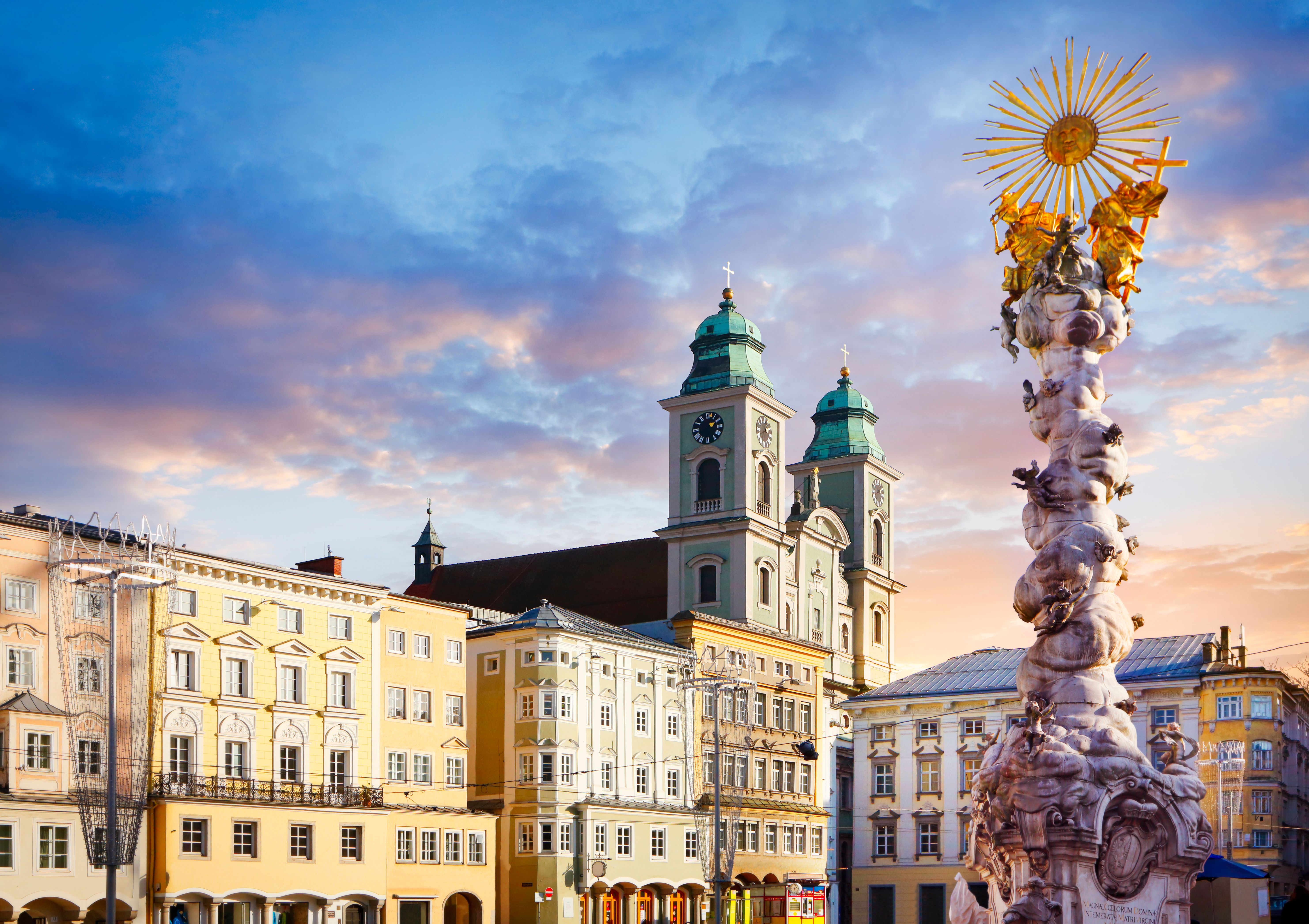 Barocke Säule mit goldenem Heiligenschein im Vordergrund, umgeben von historischen Gebäuden und einer Kirche mit zwei Türmen, bei blauem Himmel mit vereinzelten Wolken im Abendlicht.