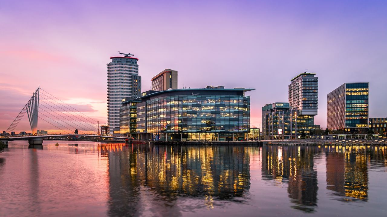 Moderne Bürogebäude am Flussufer bei Sonnenuntergang, spiegeln sich im ruhigen Wasser. Eine markante Hängebrücke verbindet die Ufer. Sanfte Abenddämmerung mit violettem und rosa Himmel schafft eine entspannte Stadtatmosphäre. Moderne Bürogebäude am Flussufer bei Sonnenuntergang, spiegeln sich im ruhigen Wasser. Eine markante Hängebrücke verbindet die Ufer. Sanfte Abenddämmerung mit violettem und rosa Himmel schafft eine entspannte Stadtatmosphäre.
