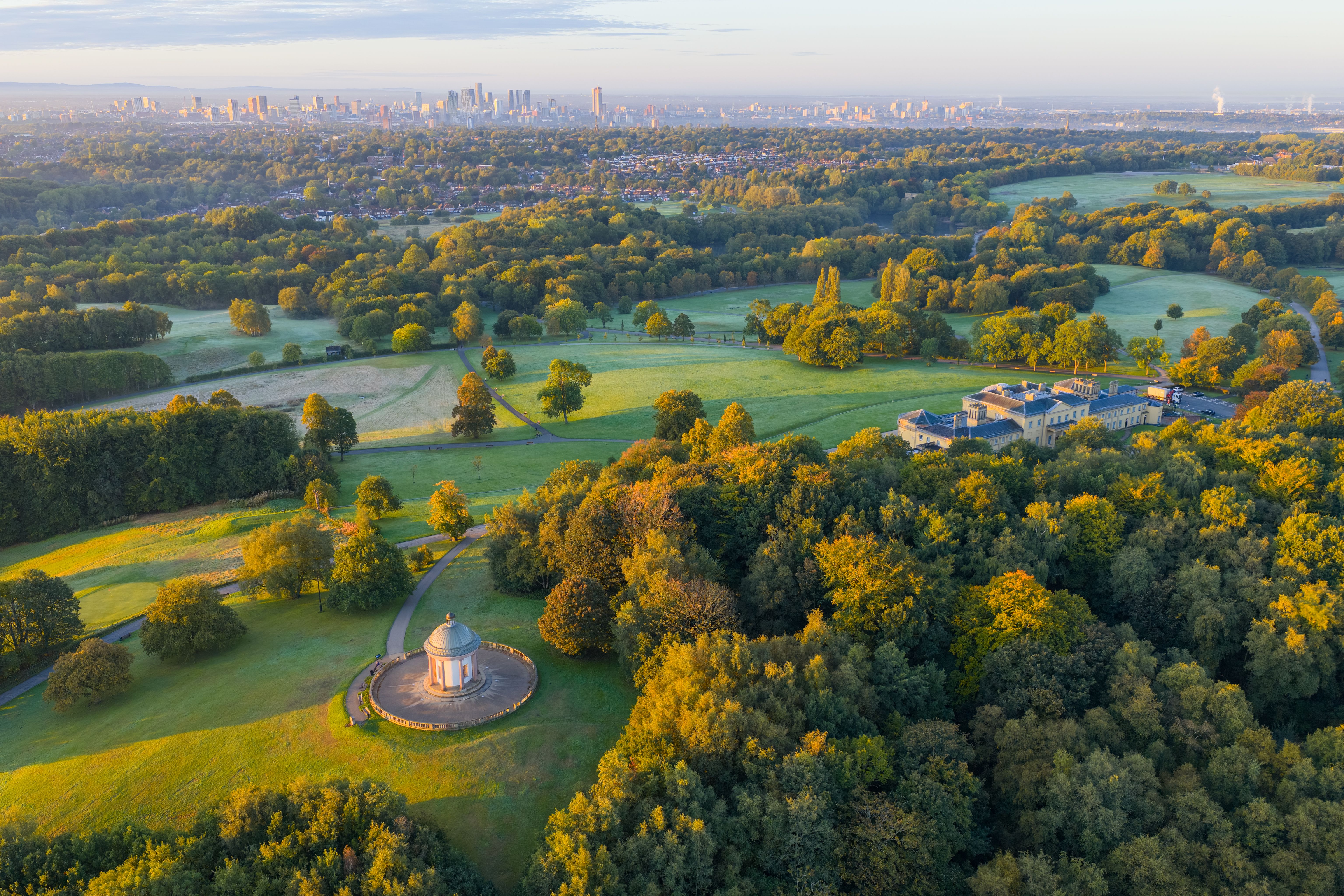 Weitblick auf einen weitläufigen Park mit grünen Wiesen und herbstlich gefärbten Bäumen, einem historischen Pavillon und einem großen Herrenhaus, im Hintergrund die Skyline einer Stadt unter blauem Himmel.
