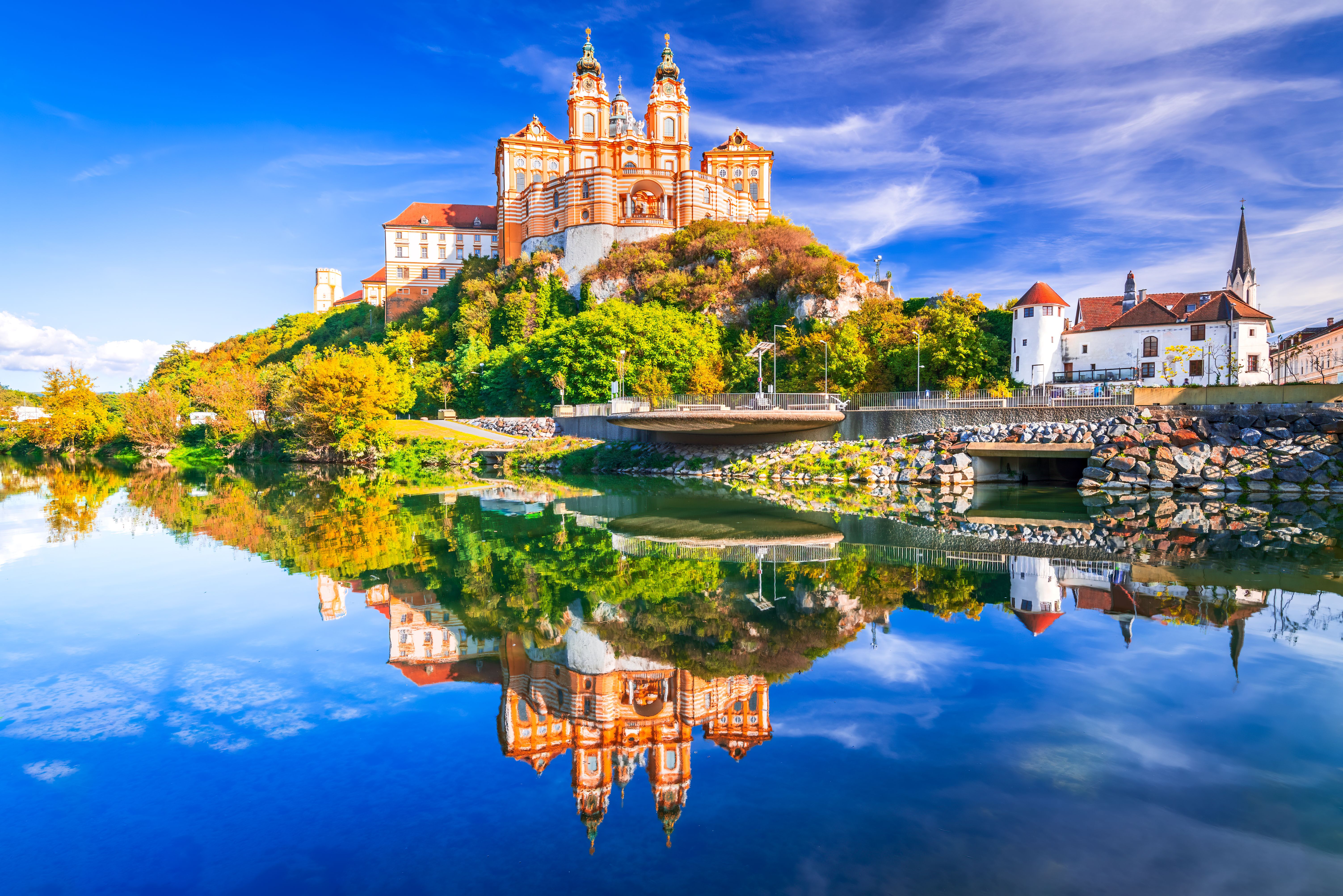 Beeindruckende Barockkirche auf einem Hügel, umgeben von grüner Natur, mit klarem, blauem Himmel und ruhigem Fluss, der das Bild spiegelglatt reflektiert – ein friedlicher und malerischer Ort für Kultur- und Naturliebhaber.