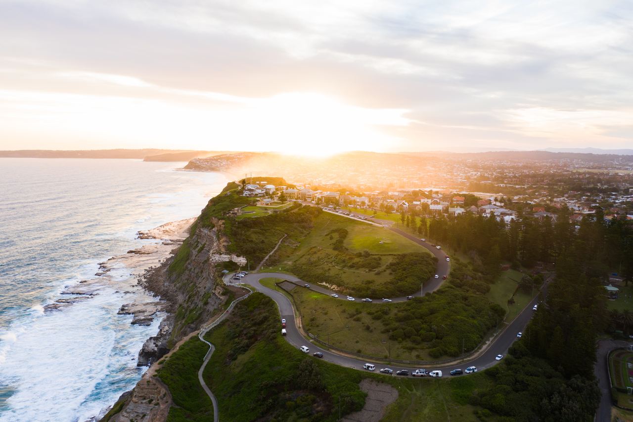Luftaufnahme einer Küstenlandschaft bei Sonnenuntergang mit einer kurvigen Straße auf einer grünen Klippe und Blick auf das Meer sowie einen Ort im Hintergrund. Die Szene vermittelt Ruhe und Weite. Luftaufnahme einer Küstenlandschaft bei Sonnenuntergang mit einer kurvigen Straße auf einer grünen Klippe und Blick auf das Meer sowie einen Ort im Hintergrund. Die Szene vermittelt Ruhe und Weite.