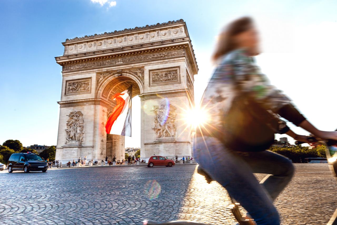 Arc de Triomphe bei Sonnenuntergang mit wehender französischer Flagge, belebte Straße und vorbeifahrende Radfahrerin, die das pulsierende Stadtleben von Paris zeigt.