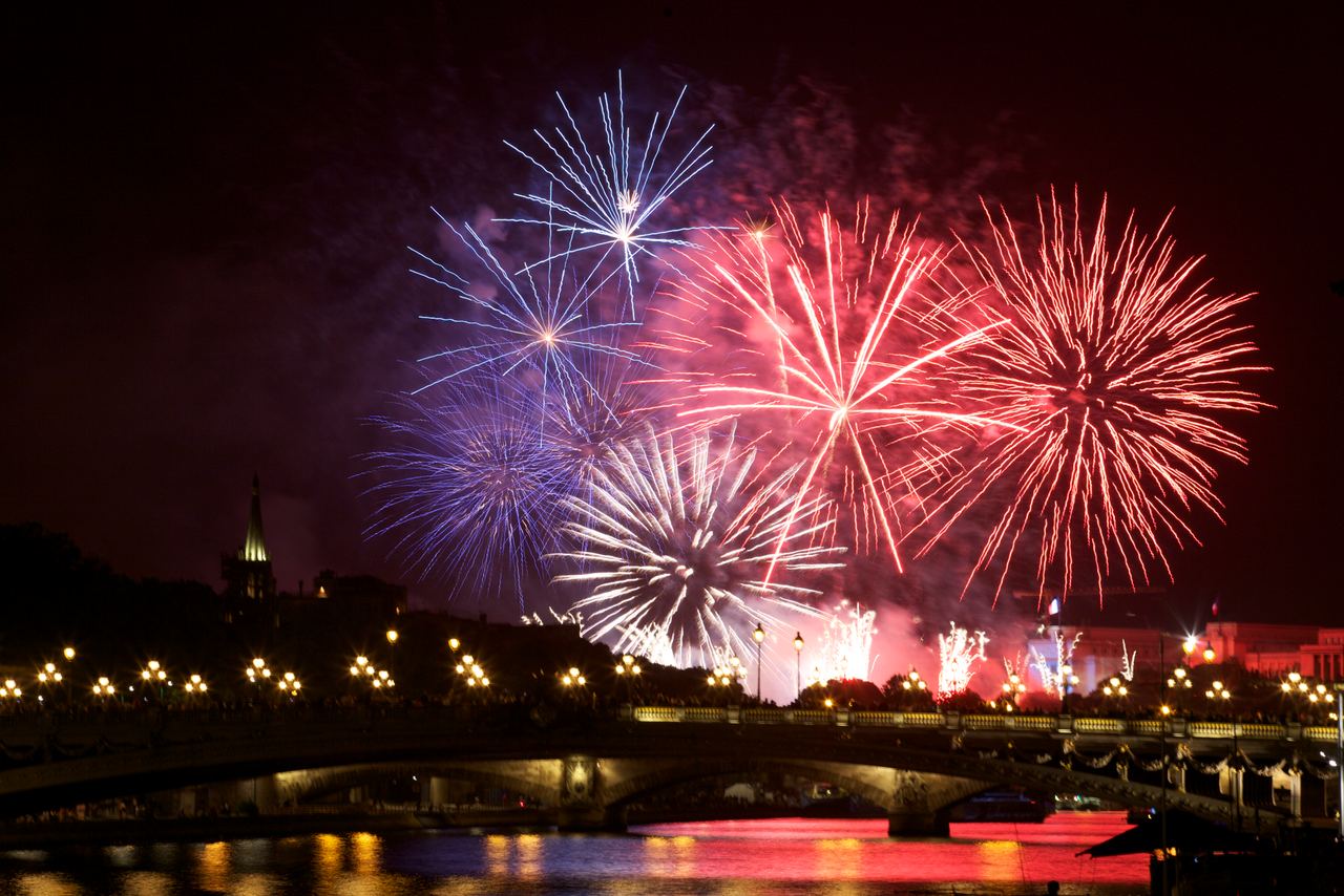 Buntes Feuerwerk erhellt die Nachthimmel über einer beleuchteten Stadtbrücke mit Reflexionen im Wasser, Feierstimmung wird vermittelt.