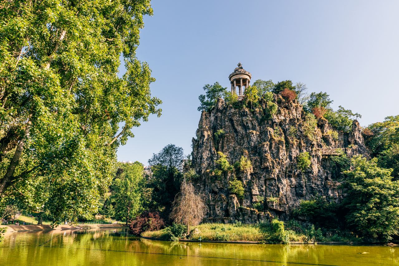 Blick auf einen felsigen Hügel mit einem Pavillon oben, umgeben von grünen Bäumen und einem ruhigen See im Vordergrund, bei klarem Himmel.