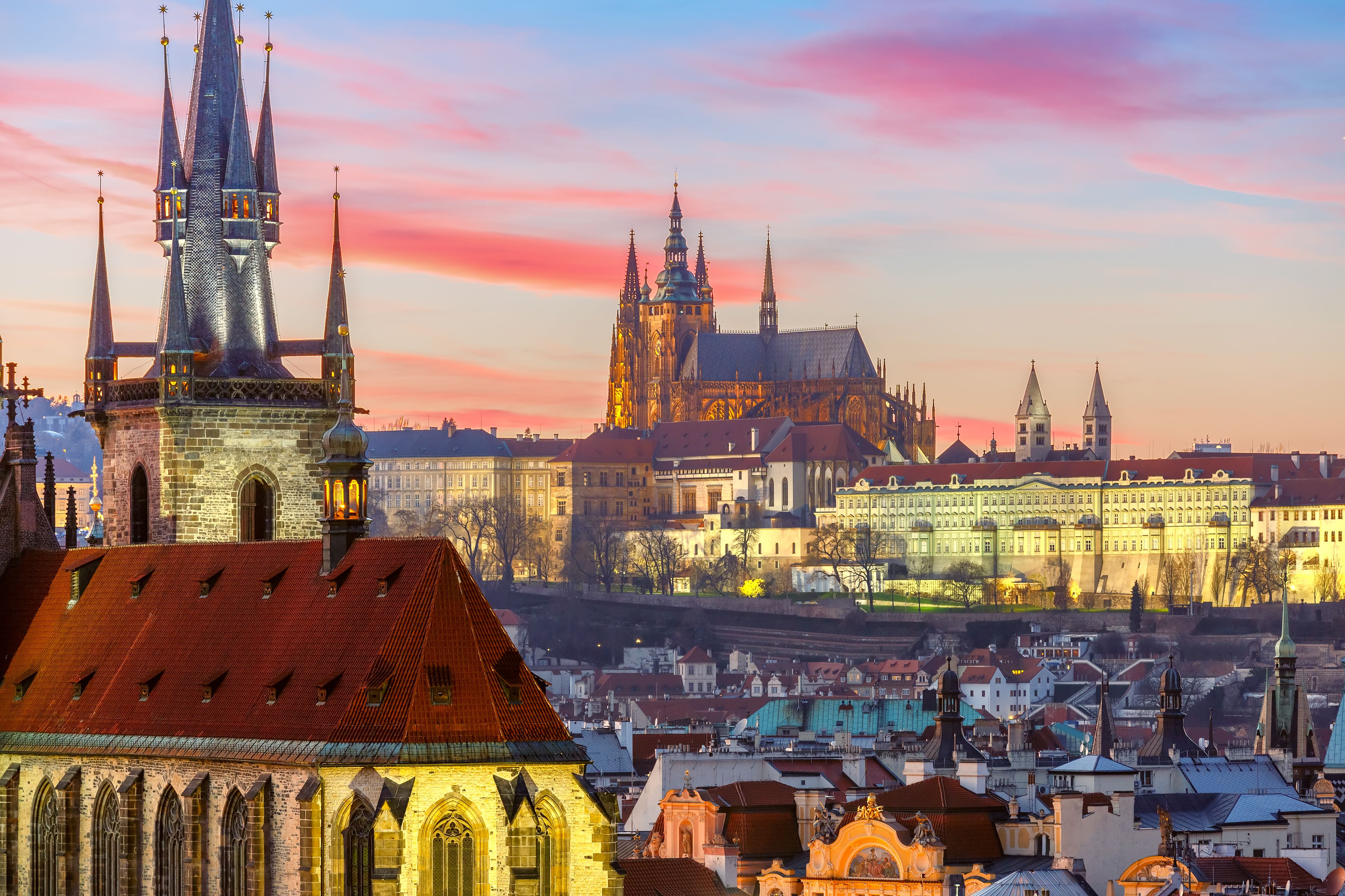 Blick auf die historische Prager Altstadt mit markanten gotischen Türmen und der beleuchteten Prager Burg im Hintergrund, unter einem malerischen rosa-blauen Abendhimmel. Ideal für Reisende, die Kultur und beeindruckende Stadtsilhouetten erleben möchten.