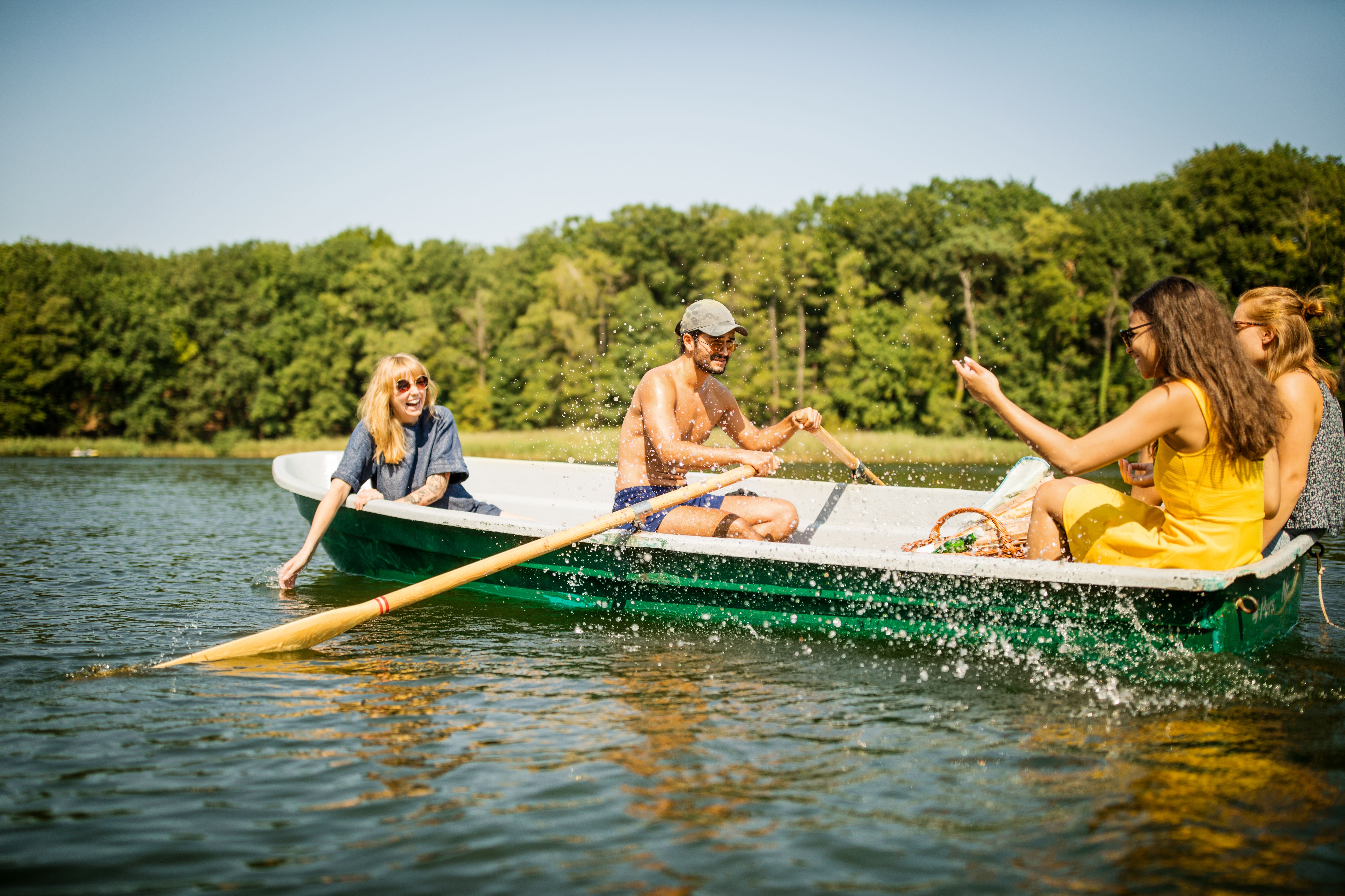 Vier Freunde genießen sonnigen Tag in einem Ruderboot auf ruhigem See, umgeben von grünen Bäumen, fröhlich und entspannt.