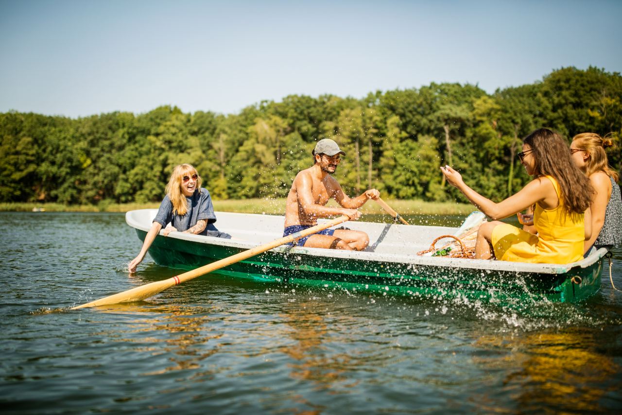 Vier Freunde genießen sonnigen Tag in einem Ruderboot auf ruhigem See, umgeben von grünen Bäumen, fröhlich und entspannt.