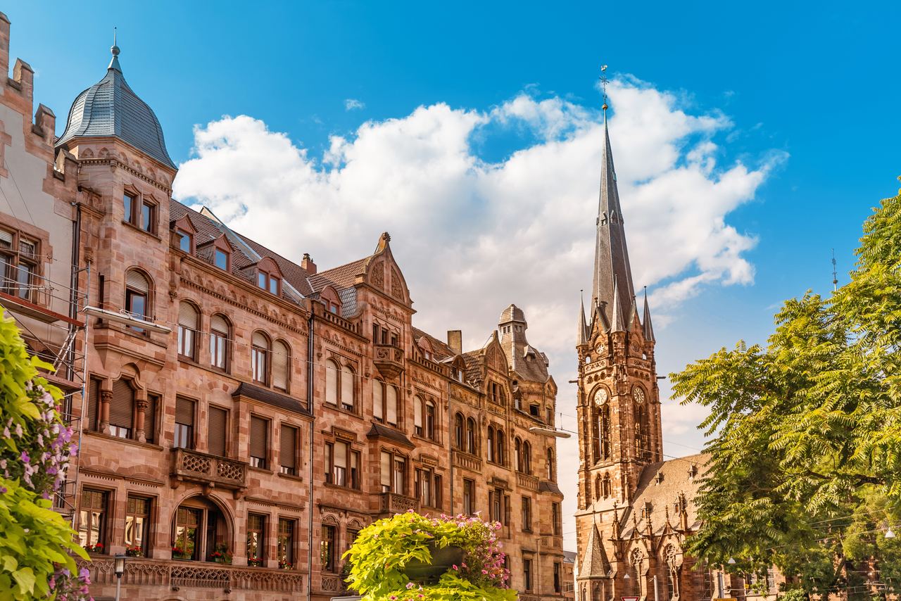 Historische Sandsteingebäude mit detailreicher Fassade und ein hoher, spitzer Kirchturm unter blauem Himmel, umgeben von grünen Bäumen, vermitteln ein charmantes Stadtbild.