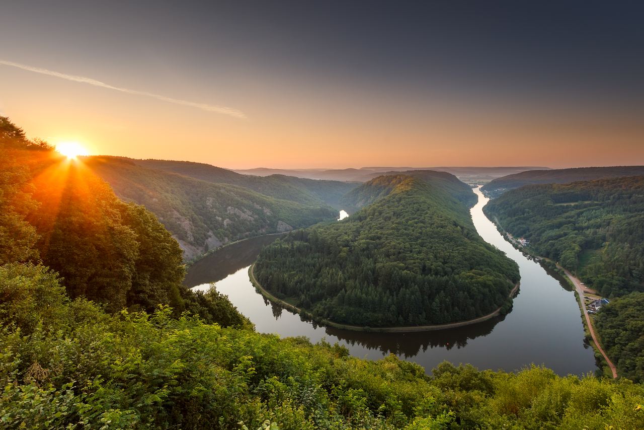 Sonnenuntergang über einer geschwungenen Flusslandschaft mit dicht bewaldeten Hügeln, die ruhige Natur und Weite ausstrahlt. Ideal für Naturliebhaber und Erholungssuchende.