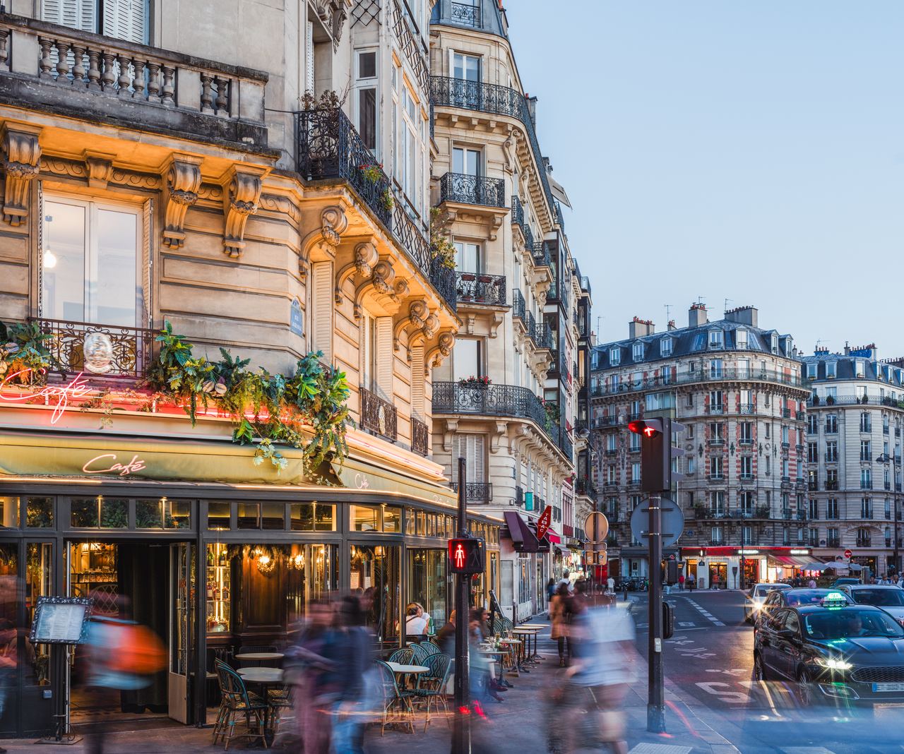 Straßenszene einer belebten Pariser Ecke mit traditionellem Café, Außenbestuhlung und historischen Gebäuden bei Dämmerung, die eine gemütliche und lebendige Atmosphäre vermittelt.