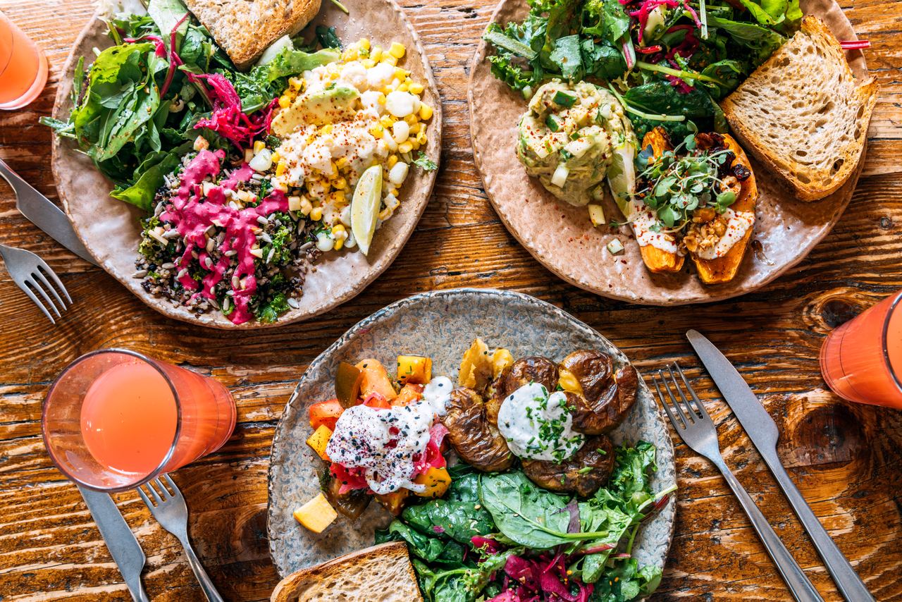 Drei Teller mit abwechslungsreichen, frischen Salaten, Gemüse, Kräutern und Vollkornbrot auf einem Holztisch, begleitet von Gläsern mit frisch gepresstem Orangensaft.