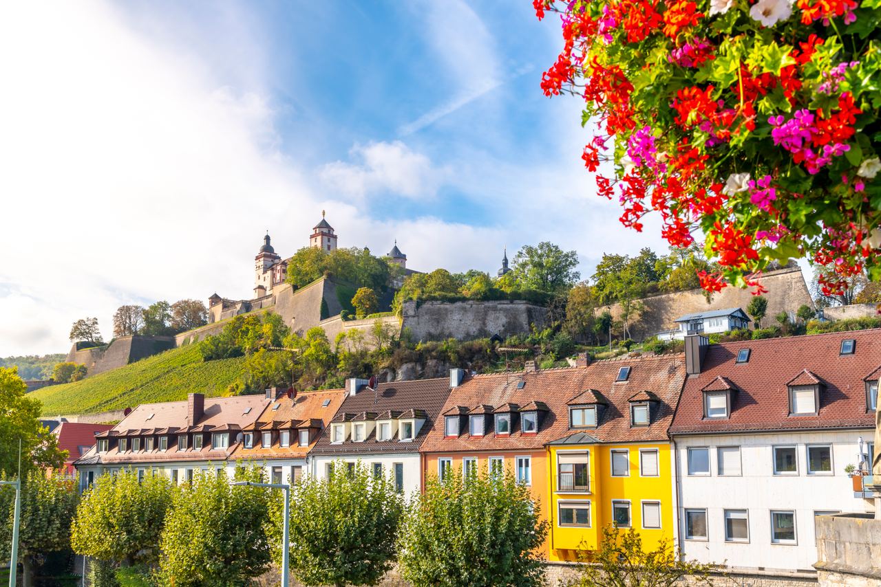 Blick auf farbenfrohe Häuserreihen vor einer grünen Anhöhe mit historischer Festung unter blauem Himmel, dekoriert mit bunten Blumen im Vordergrund.