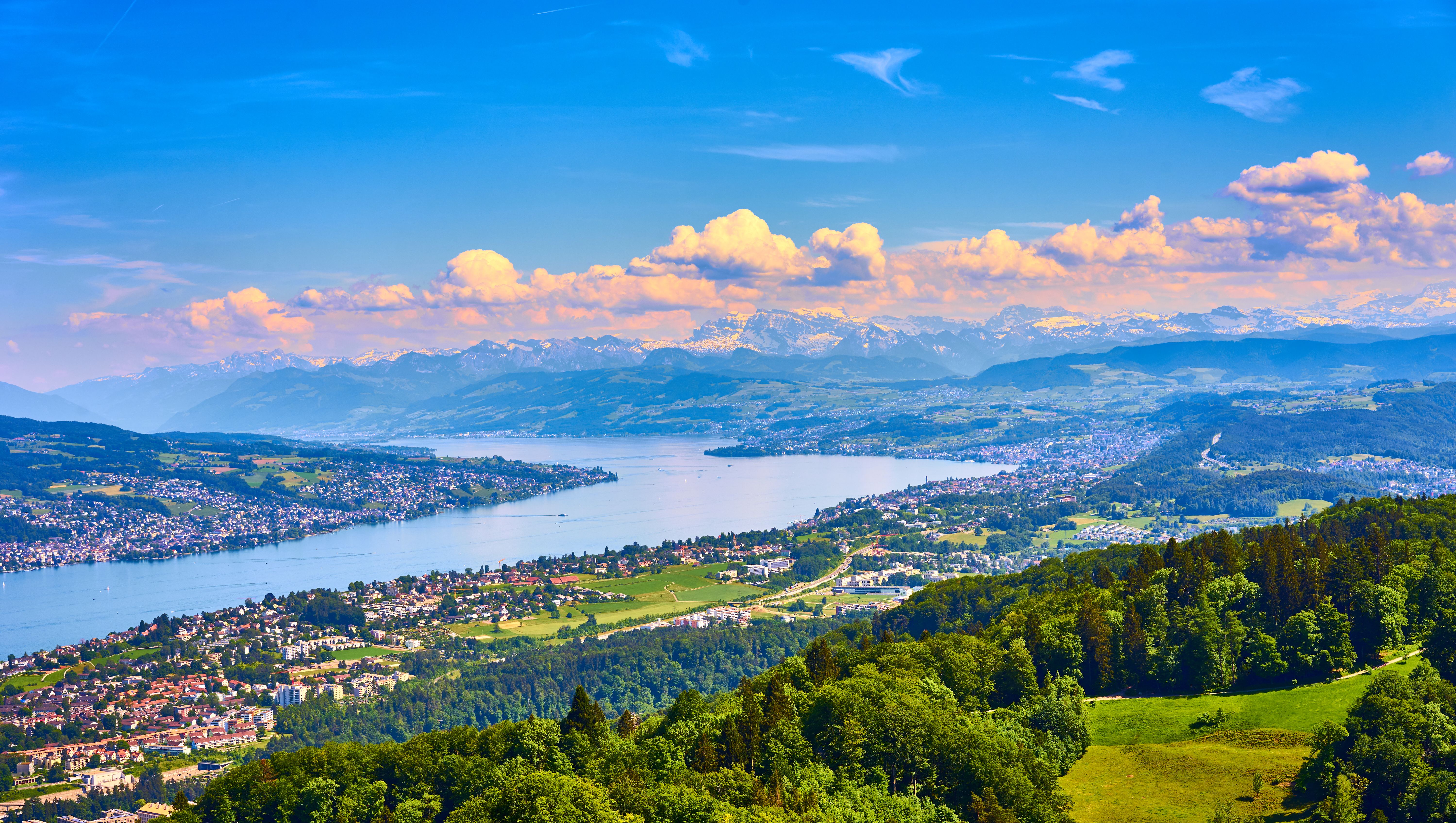 Weitläufige Panoramaansicht auf einen großen See, umgeben von grünen Hügeln und Städten, im Hintergrund majestätische Berge unter blauem Himmel mit weißen Wolken.