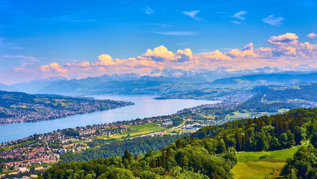 Weitläufige Panoramaansicht auf einen großen See, umgeben von grünen Hügeln und Städten, im Hintergrund majestätische Berge unter blauem Himmel mit weißen Wolken. Weitläufige Panoramaansicht auf einen großen See, umgeben von grünen Hügeln und Städten, im Hintergrund majestätische Berge unter blauem Himmel mit weißen Wolken.