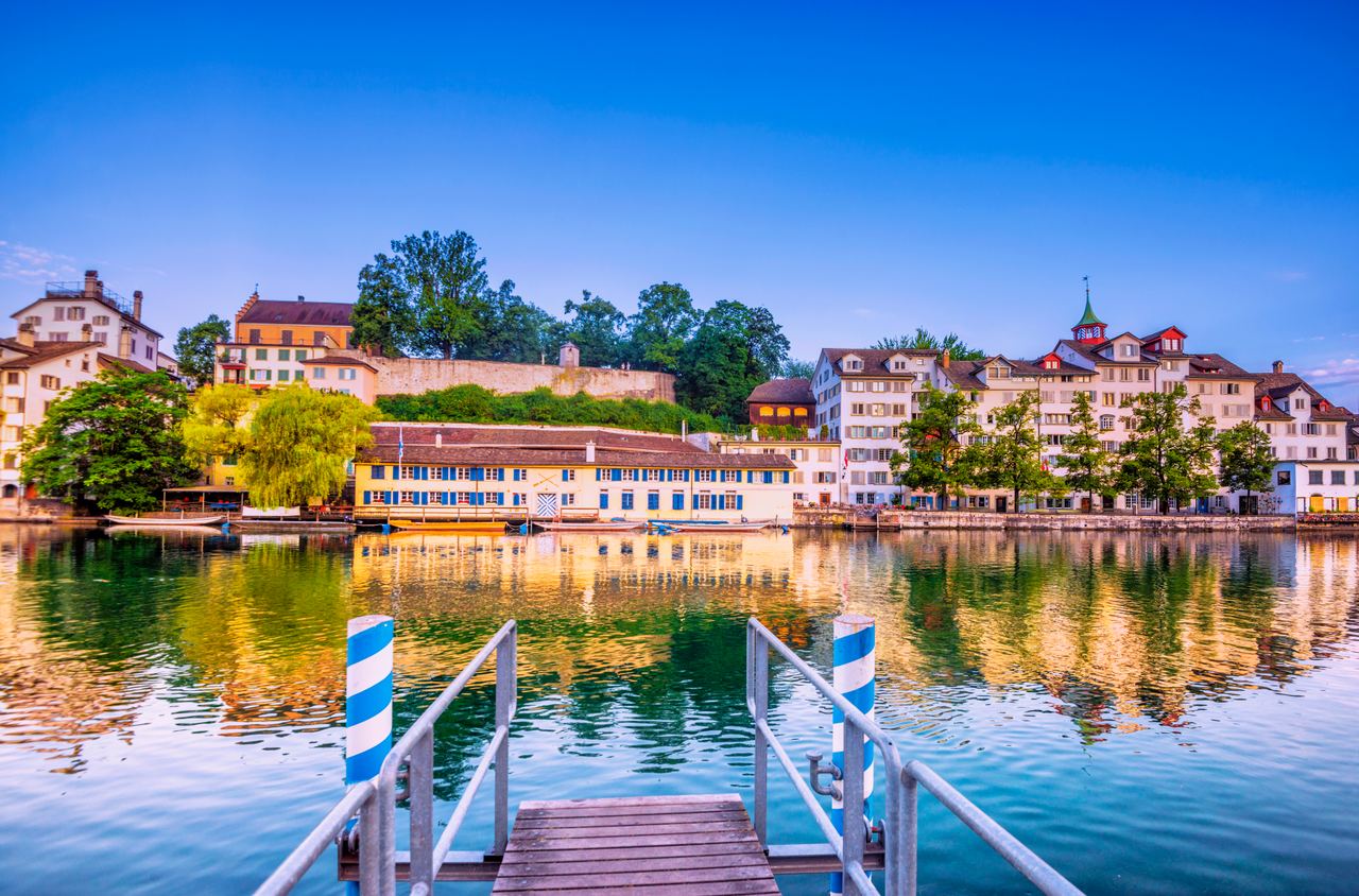 Uferpromenade mit historischen Gebäuden und grünen Bäumen spiegelt sich ruhig im Wasser. Im Vordergrund ein kleiner Steg mit weißen und blauen Pfosten lädt zum Verweilen ein. Klare, blaue Abendstimmung über der Stadt am Fluss.