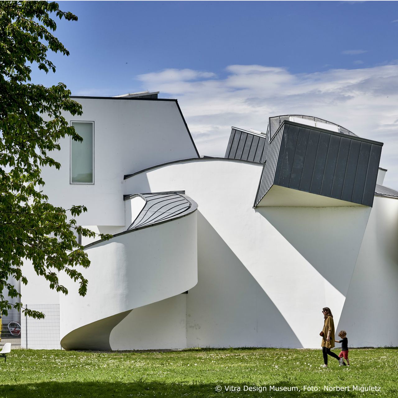 Weißes, modernes Museum mit geschwungenen und eckigen Architekturformen, umgeben von Grünfläche; Frau mit Kind spaziert davor.