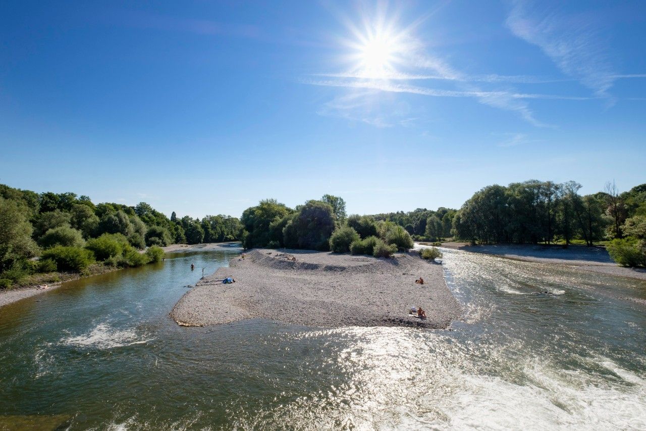 Fluss mit einer kleinen Kiesinsel in der Mitte, umgeben von grünem Wald, unter strahlend blauem Himmel mit heller Sonne. Einige Menschen entspannen auf der Insel.