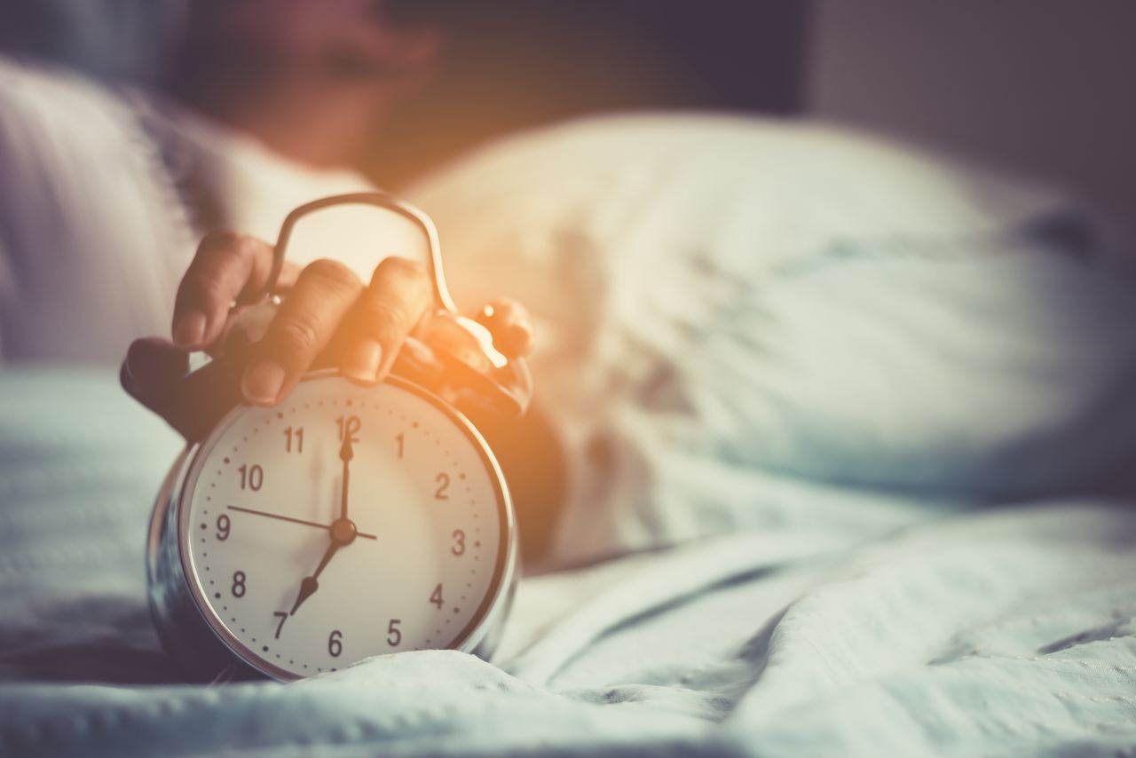 Hand, die einen klassischen Wecker auf einem Bett berührt, Symbol für Morgenroutine und Aufwachen im Hotel.