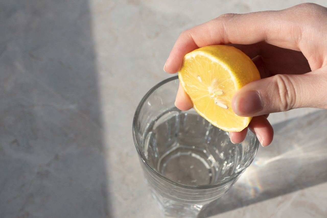Hand drückt eine Zitronenhälfte über einem Glas mit klarem Wasser, um frischen Zitronensaft hinzuzufügen.