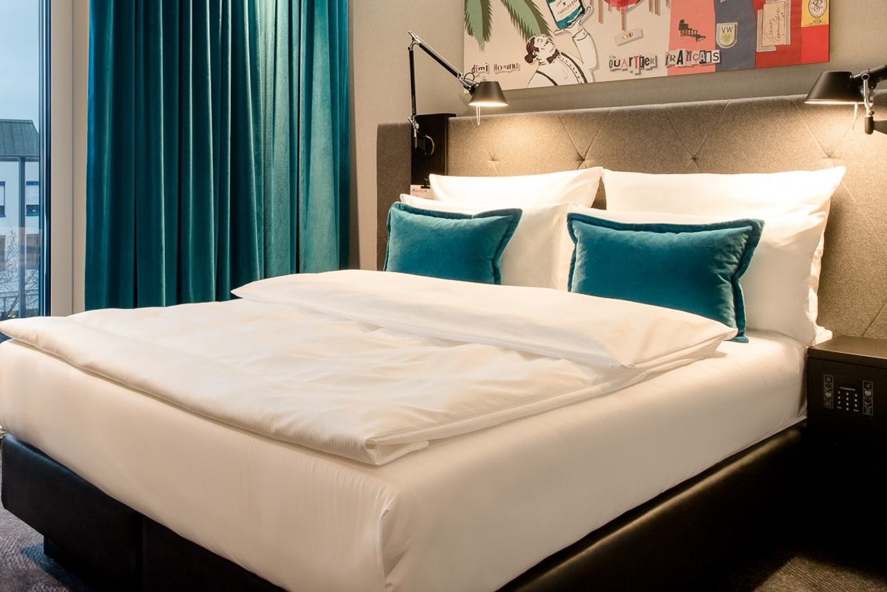 Hotelzimmer mit großem, gemütlichem Doppelbett, weißen Bettdecken und türkisfarbenen Kissen, stimmungsvolle Beleuchtung und Blick auf eine Stadt durch das Fenster.