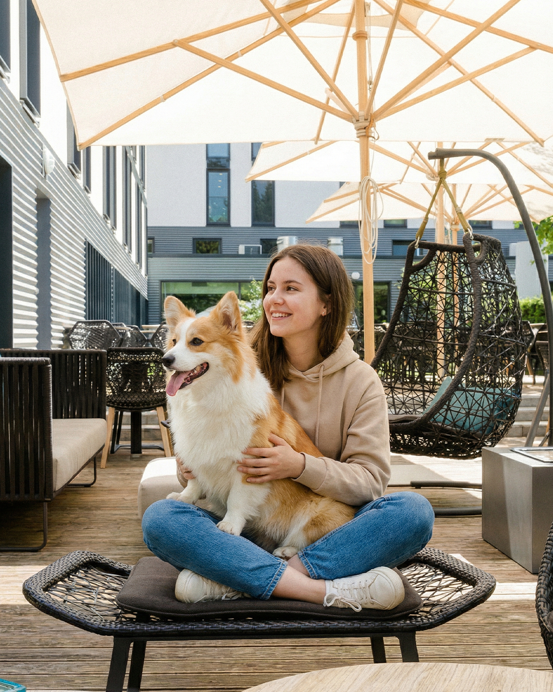 Junge Frau sitzt entspannt auf Terrasse unter Sonnenschirmen, hält glücklich einen Hund auf dem Schoß. Moderne Außenmöbel und freundliche Atmosphäre laden zum Verweilen ein.