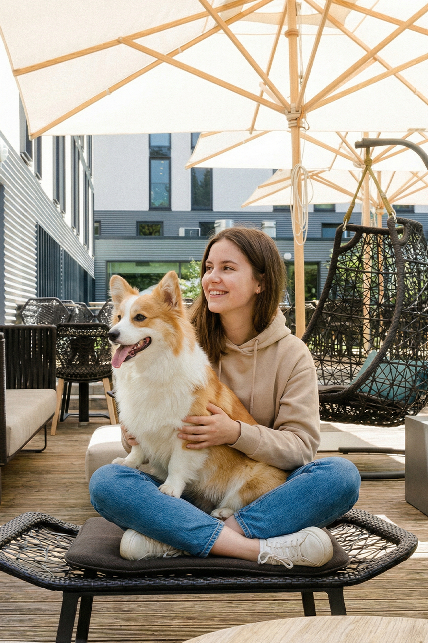 Junge Frau sitzt entspannt auf Terrasse unter Sonnenschirmen, hält glücklich einen Hund auf dem Schoß. Moderne Außenmöbel und freundliche Atmosphäre laden zum Verweilen ein.