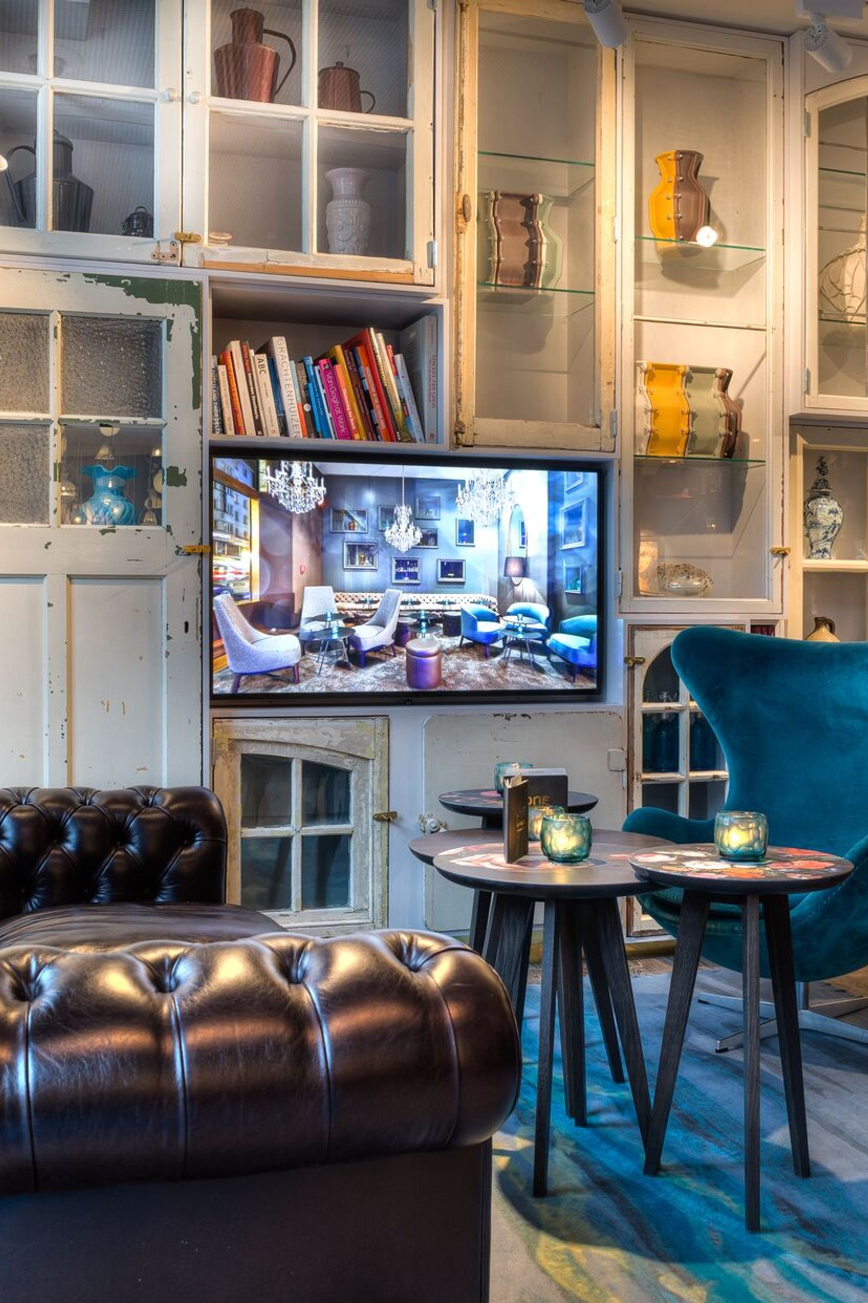 Lounge-Bereich mit dunkelbraunem Leder-Sofa, Samt-Sessel in Blau, kleinen Tischen und Bücherregal in hellem Vintage-Stil