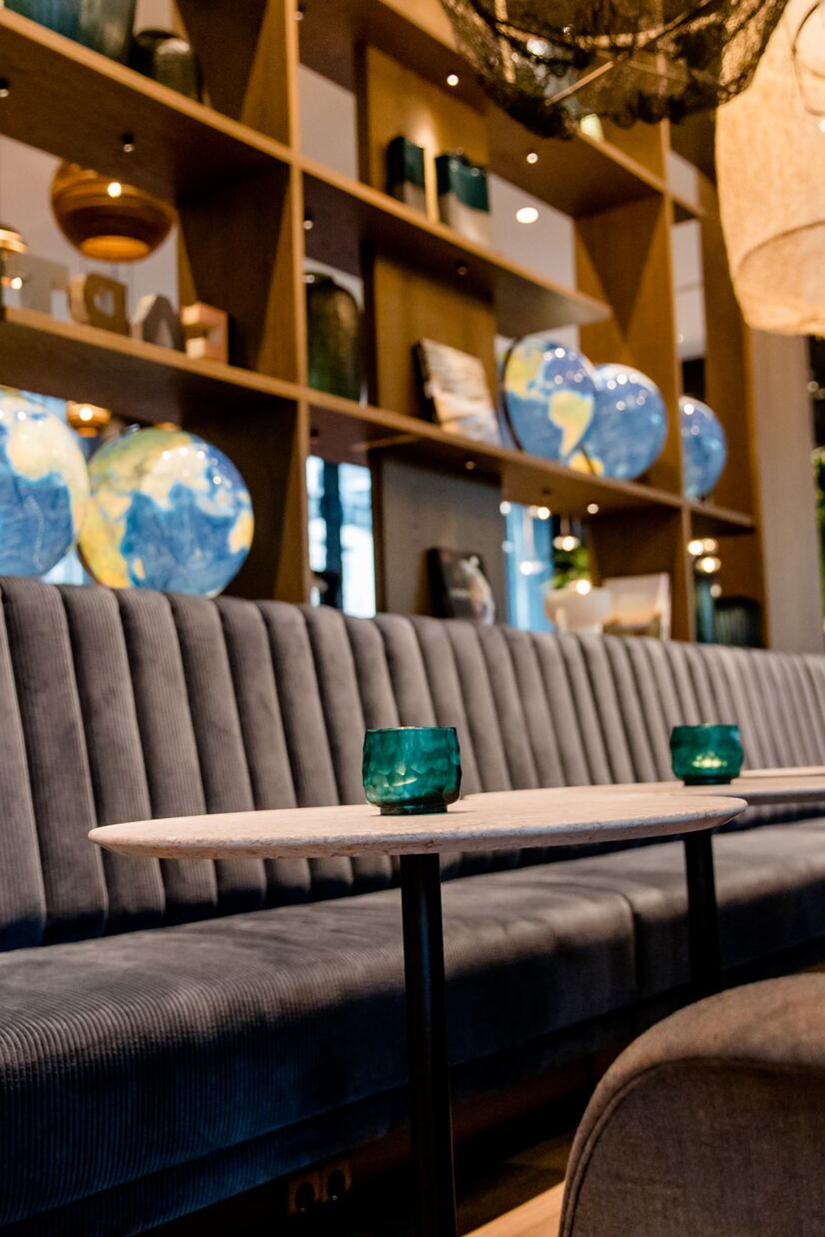 Hotel-Lounge mit gepolsterten Sitzbänken, kleinen Tischen und stimmungsvollem Licht, umgeben von dekorativen Weltkugeln und offenen Regalen.