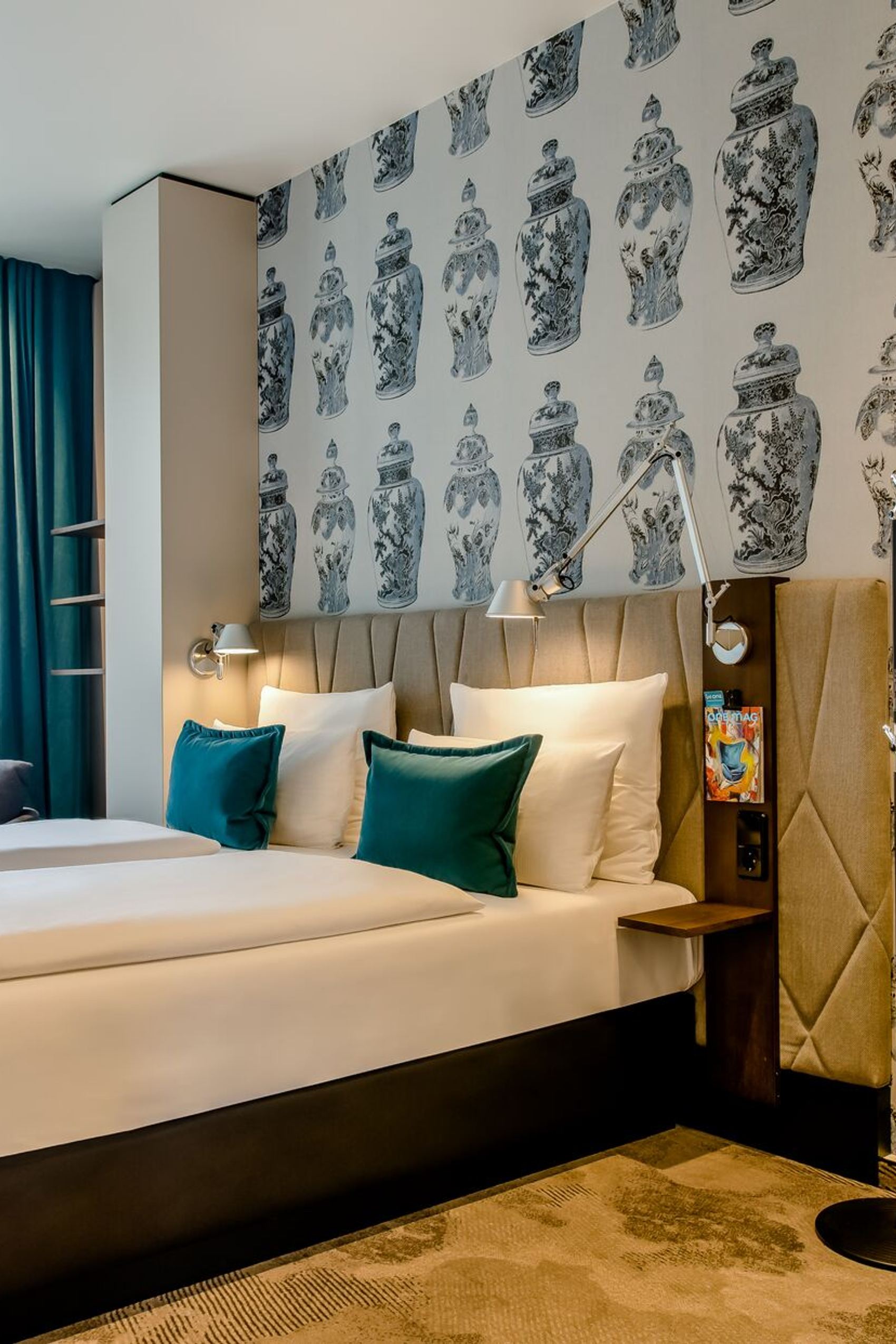 Hotelzimmer mit zwei Einzelbetten, bequemen Kissen, hellen Wänden und dekorativer Tapete mit Vasenmotiven. Große Fenster bieten Tageslicht und einen Blick auf die Stadt. Gemütliche Atmosphäre mit funktionaler Beleuchtung für Erholung und Arbeit.