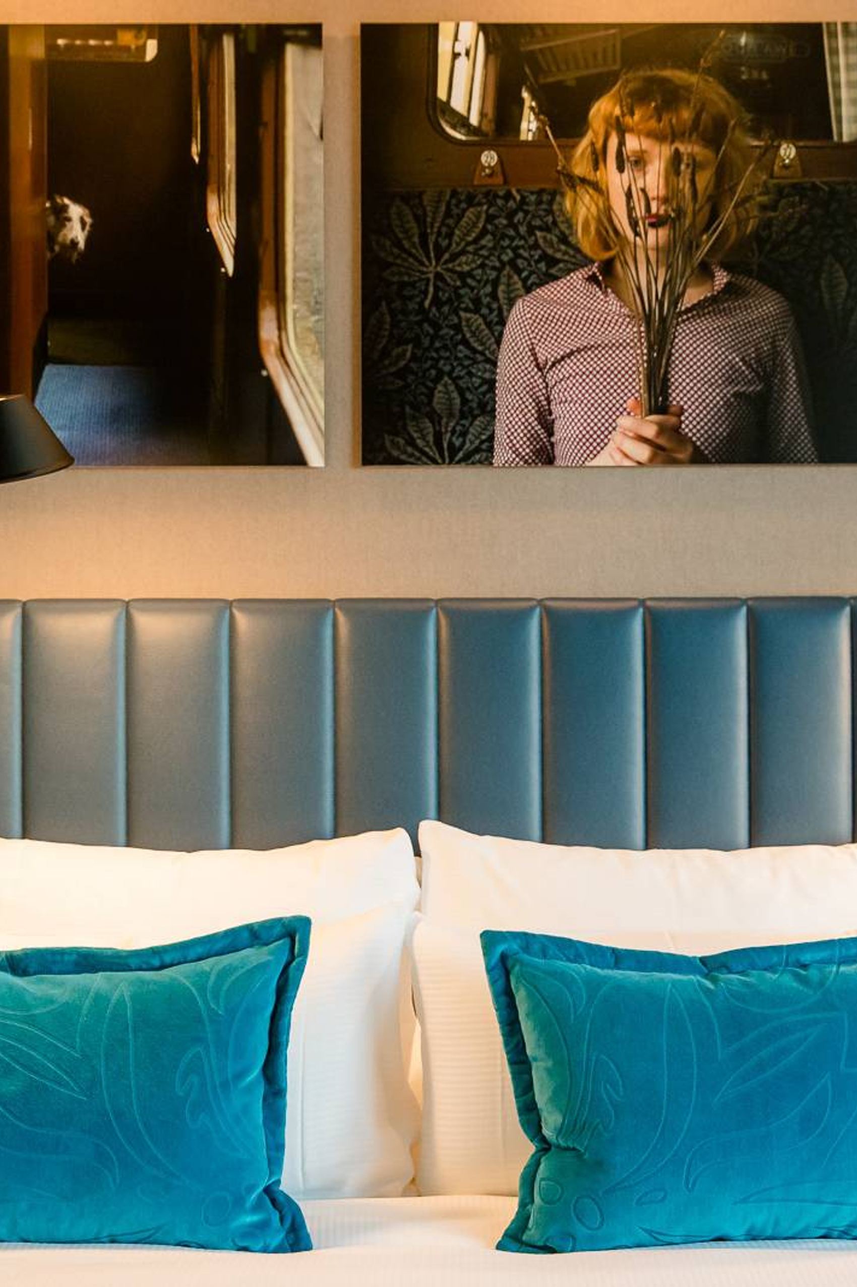 Hotelzimmer mit komfortablem Bett, blauen Samtkissen und
Wanddekoration, die eine künstlerische Fotografie einer Person zeigt.