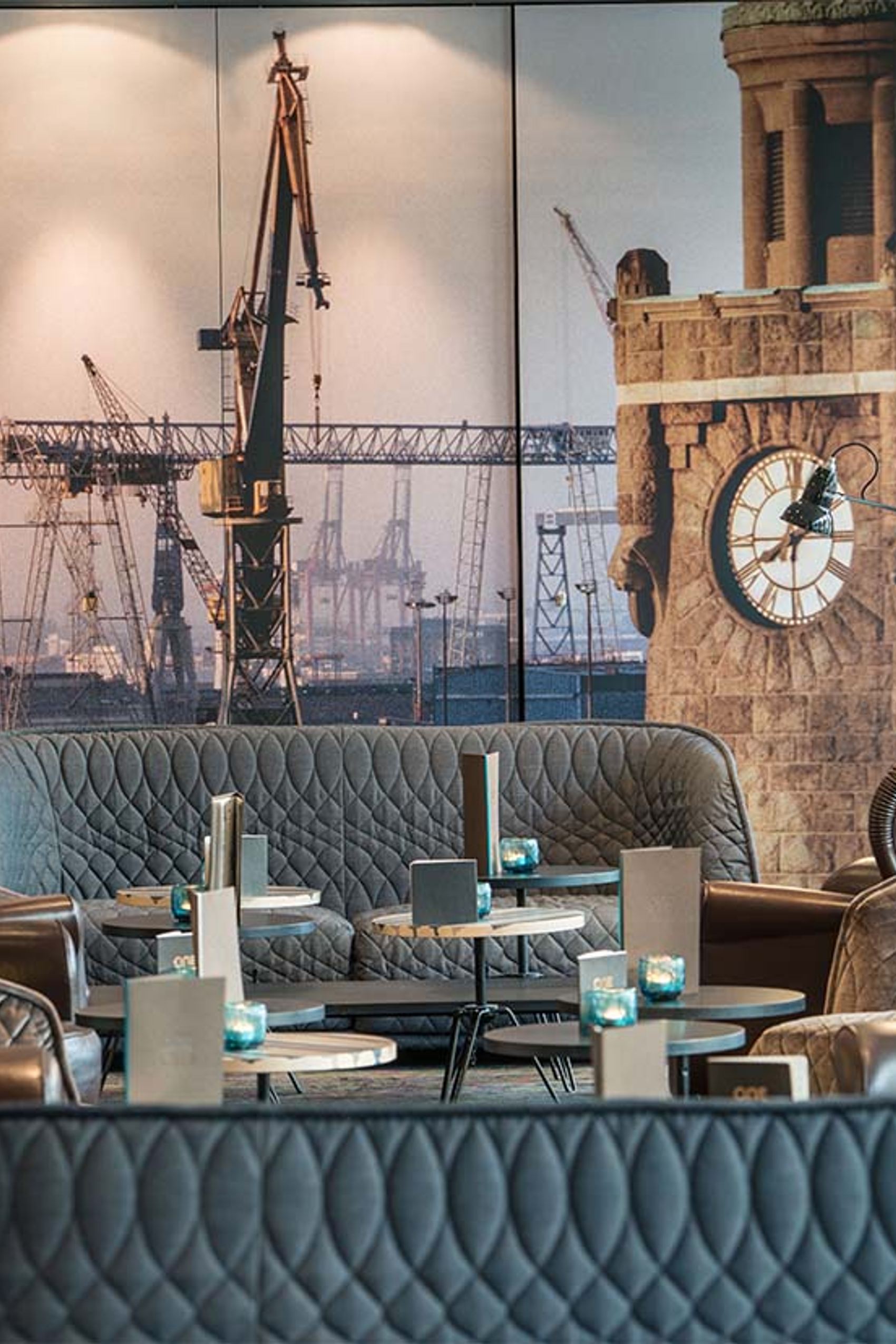 Lounge-Bereich mit ledernen Sesseln und Sofas, kleinen runden Tischen und Kerzenlicht, davor eine große Wanddekoration mit Hafenkränen und einem alten Uhrenturm.