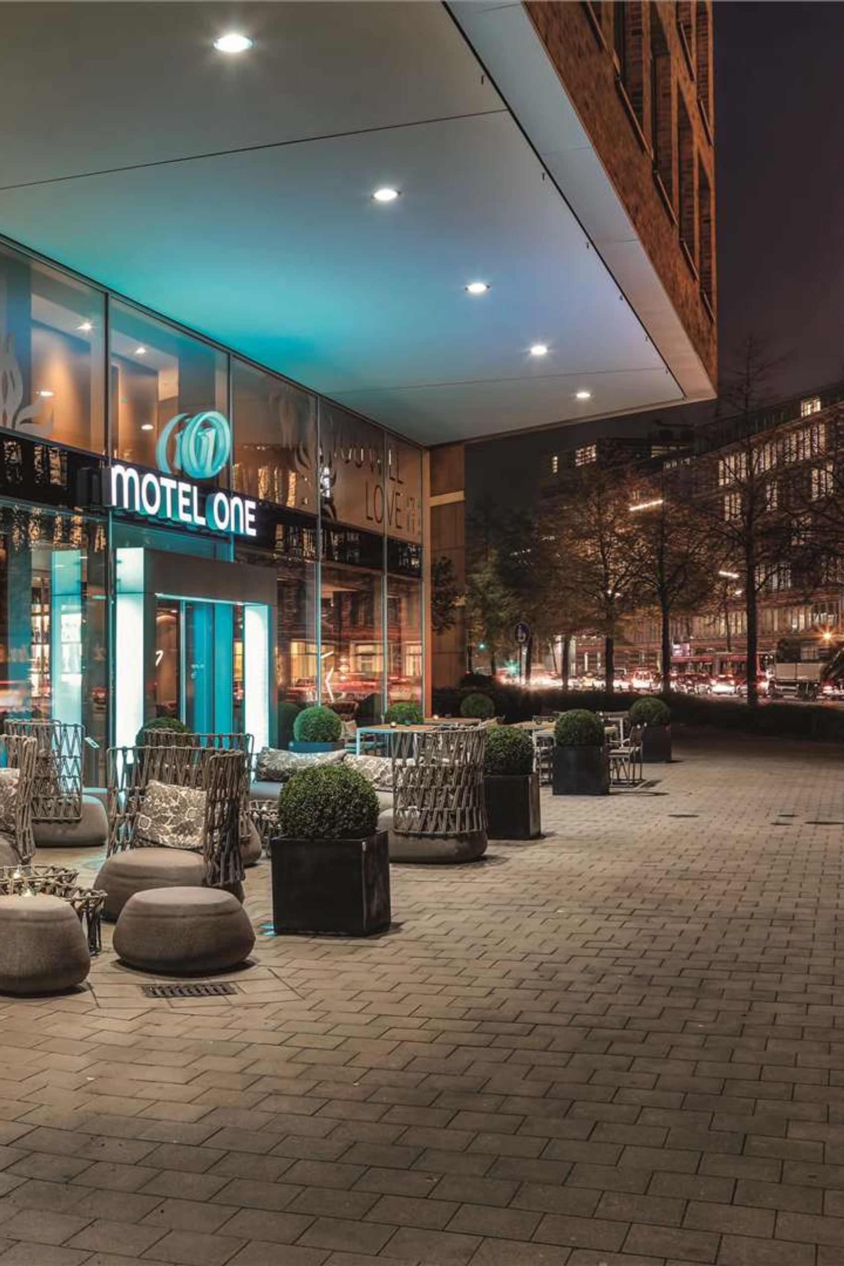 Außenansicht von Motel One bei Nacht mit modern gestalteter Terrasse, gemütlichen Sitzgelegenheiten und stimmungsvoller Beleuchtung, in urbaner Umgebung nahe historischer Gebäude.