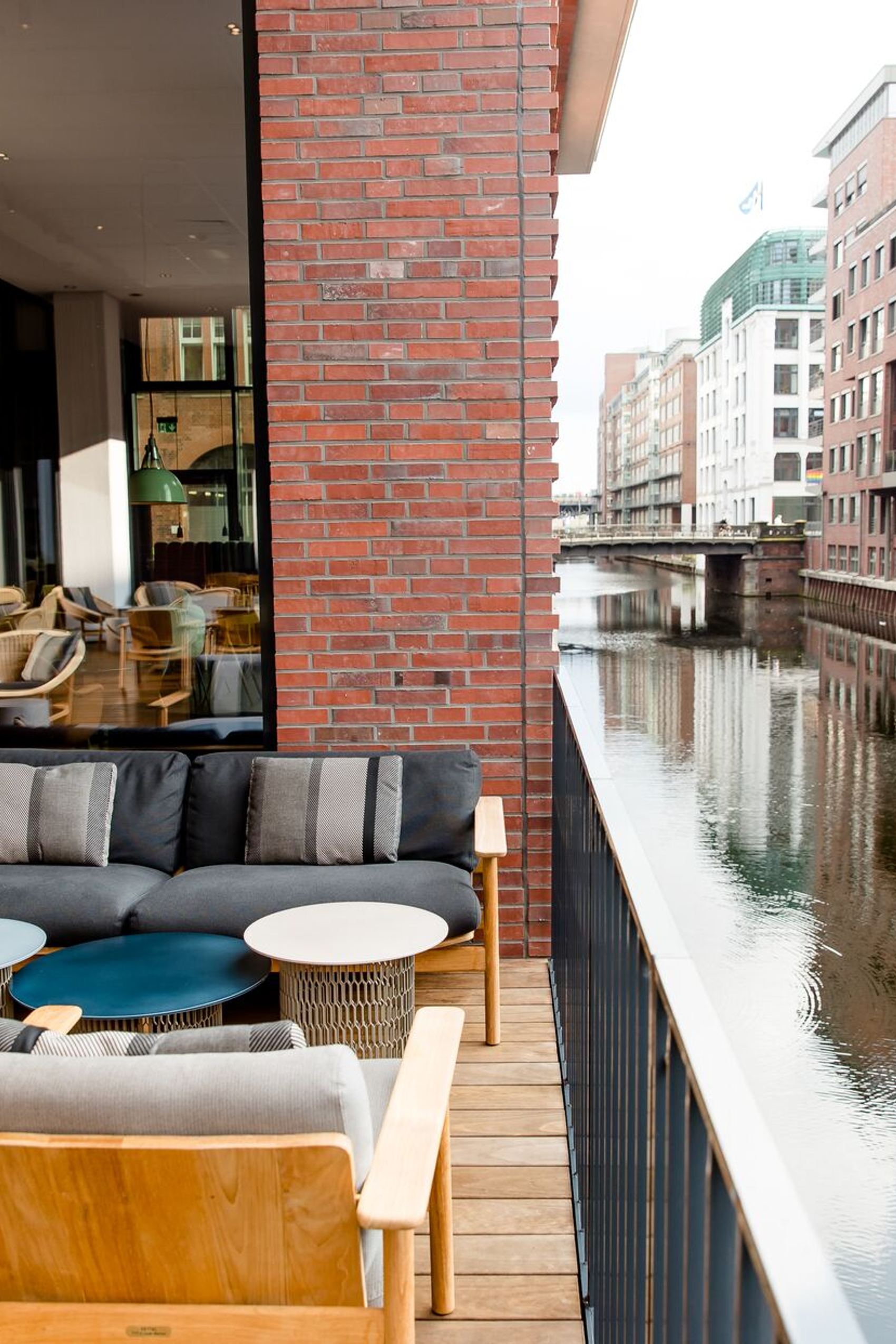 Außenbereich mit gemütlichen Sitzmöbeln auf Holzterrasse, Blick auf ruhigen Wasserkanal und moderne Backstein- und Glasgebäude, einladender Ort zum Entspannen mit urbanem Flair.