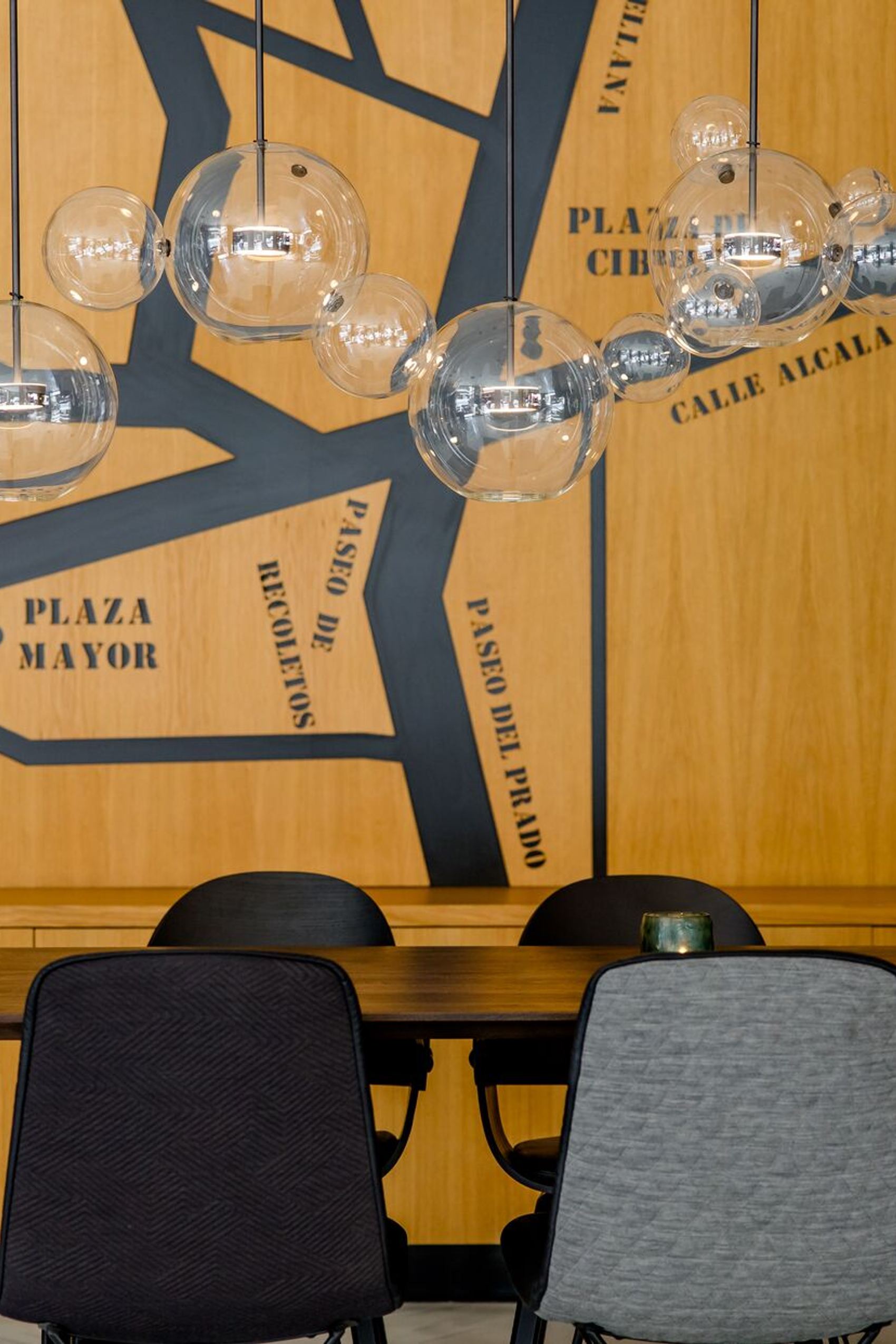Sitzbereich mit eleganten Stühlen und einem Holztisch. Im Hintergrund eine dekorative Wand mit stilisierter Stadtkarte, darüber hängen kugelförmige Glasleuchten, die für warmes, einladendes Licht sorgen.
