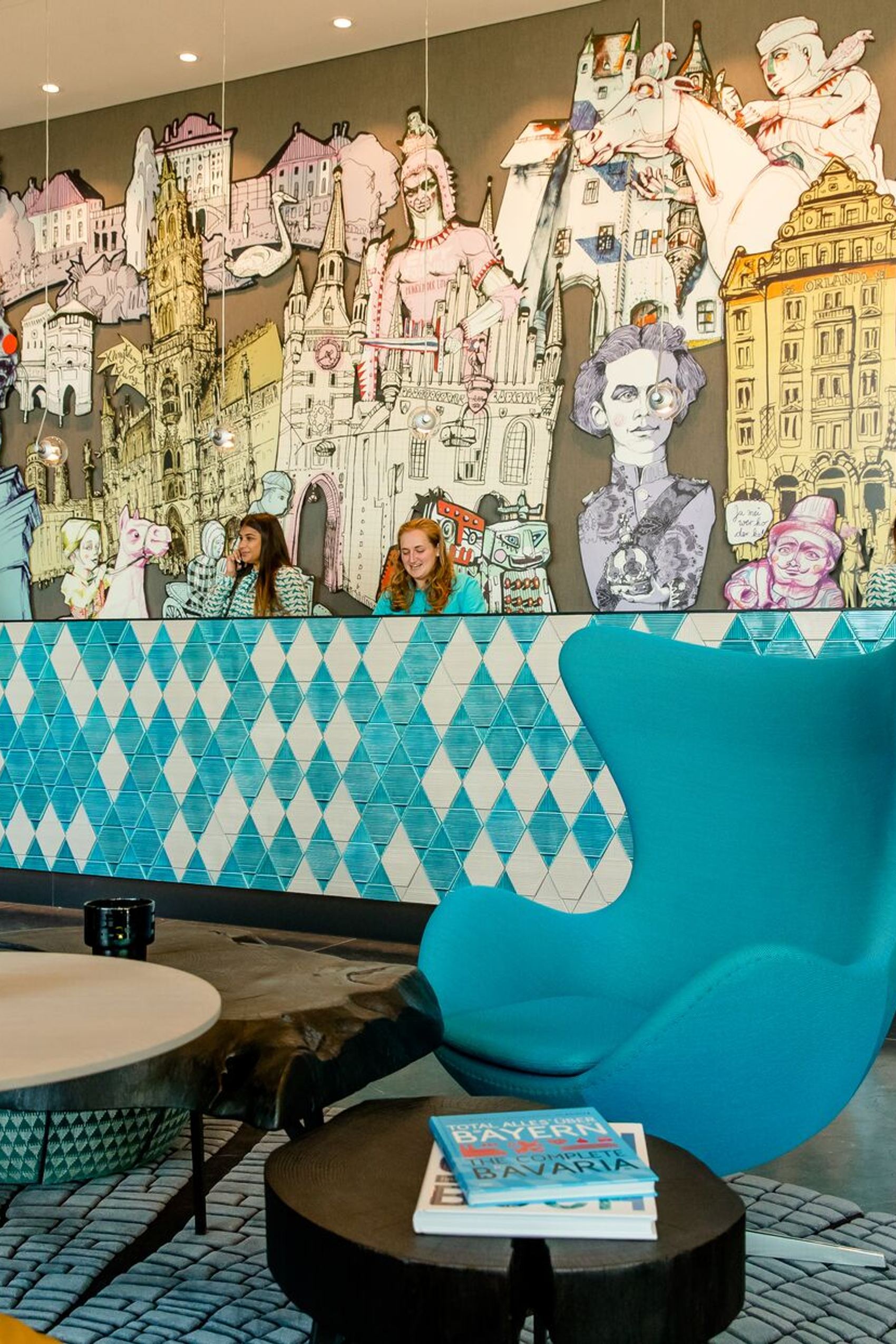Hotel-Lobby mit modernem, blau gemustertem Empfangstresen und künstlerischer Wandgestaltung, die historische Figuren und Architektur zeigt. Komfortabler blauer Sessel und Tische.