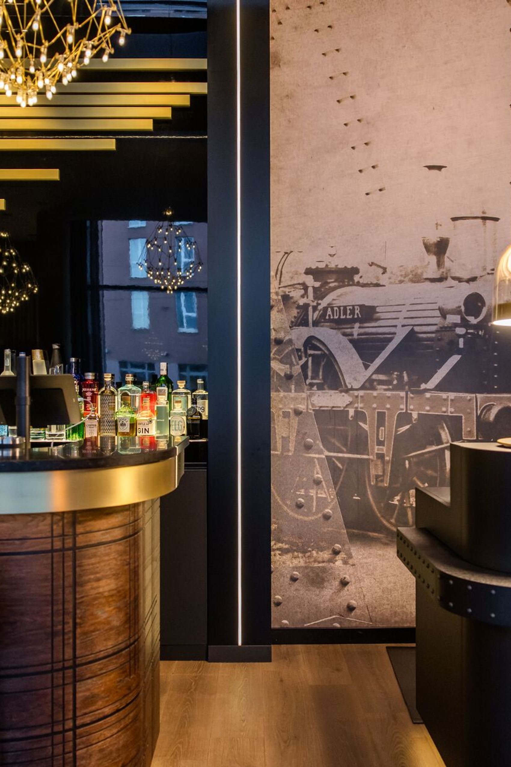 Hotelbar mit elegantem Holztresen, gut bestückter Spirituosenauswahl und stilvoller Beleuchtung. An der Wand ein großes schwarz-weiß Bild einer historischen Dampflok, das der Atmosphäre einen besonderen Charakter verleiht.
