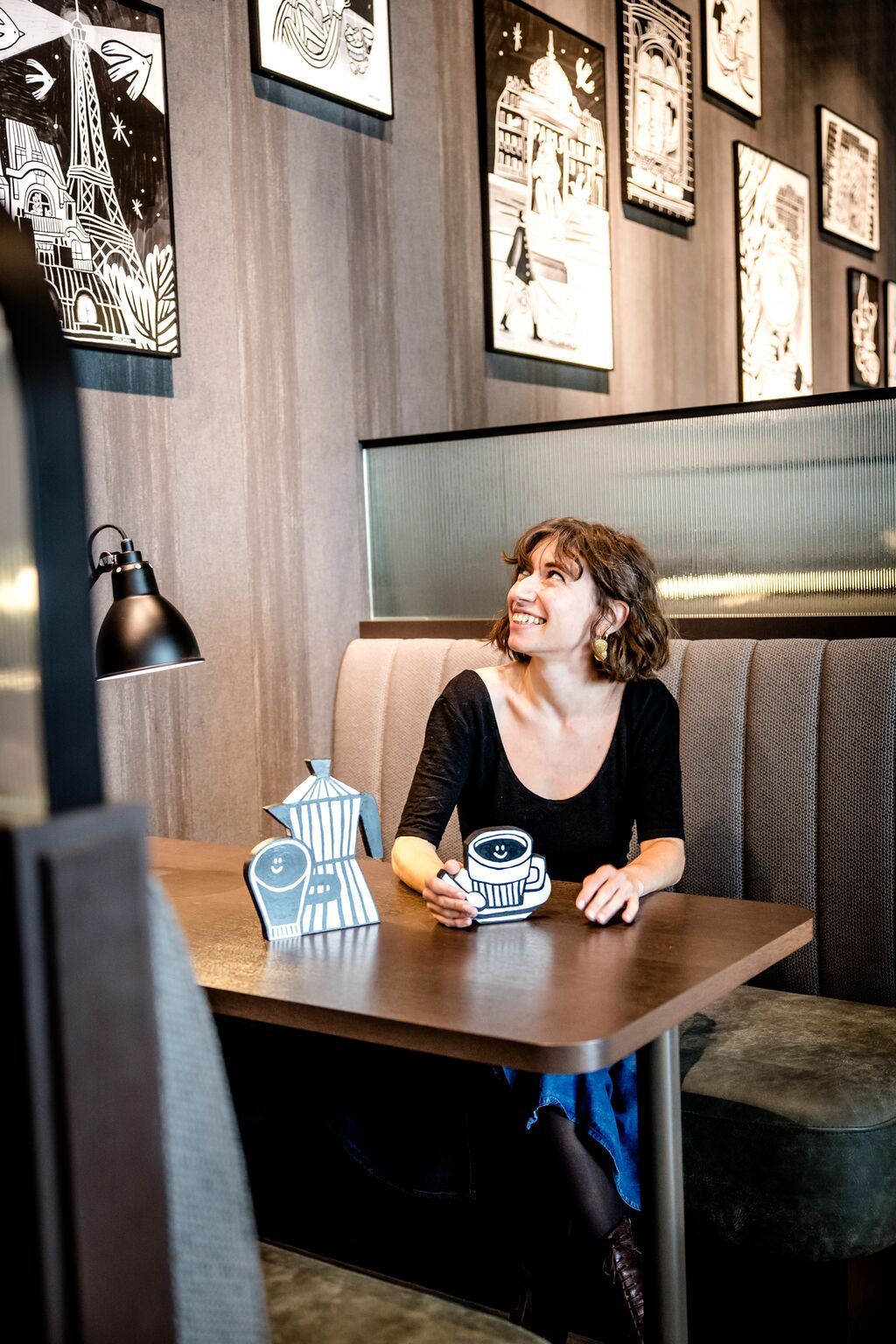 Frau sitzt entspannt in einer stilvollen Café-Ecke, hält eine Tasse und lächelt, umgeben von dekorativen Wandbildern mit Pariser Motiven.