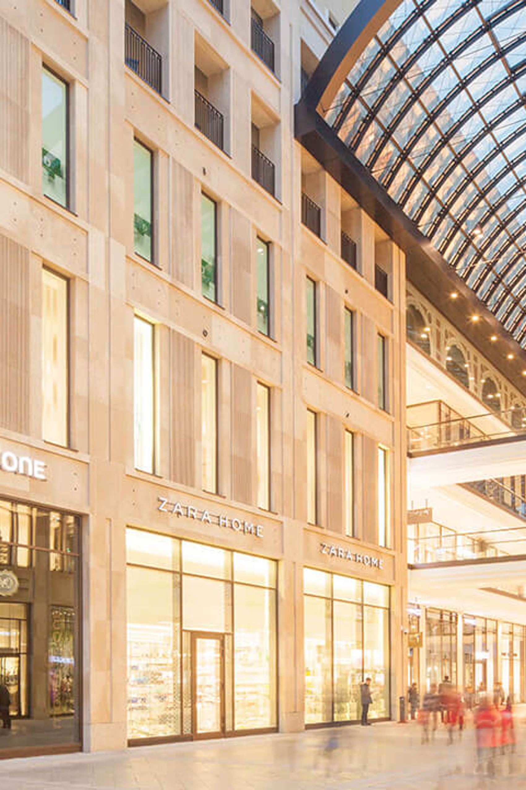 Einkaufszentrum mit großen Glasfenstern und gewölbtem Glasdach, darunter weitläufige Promenade mit Geschäften, Cafés und Spaziergängern