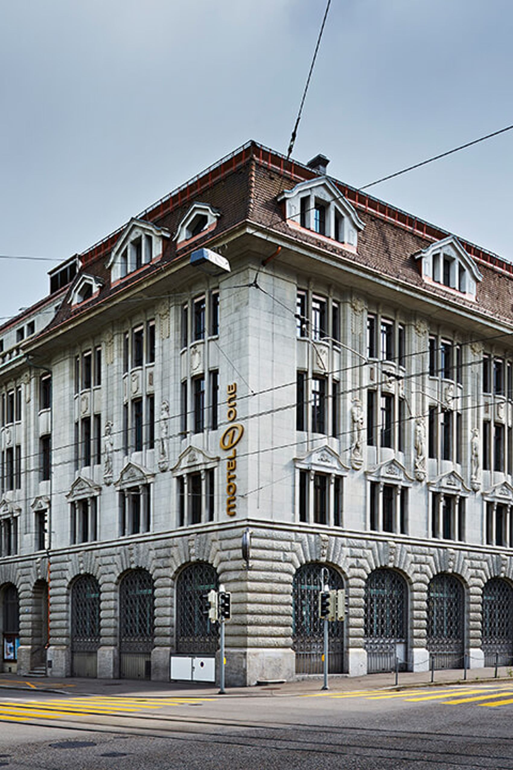 Außenansicht eines großen, historischen Gebäudes mit mehreren Stockwerken, hohem Dach und vielen Fenstern an einer Straßenecke in der Stadt.