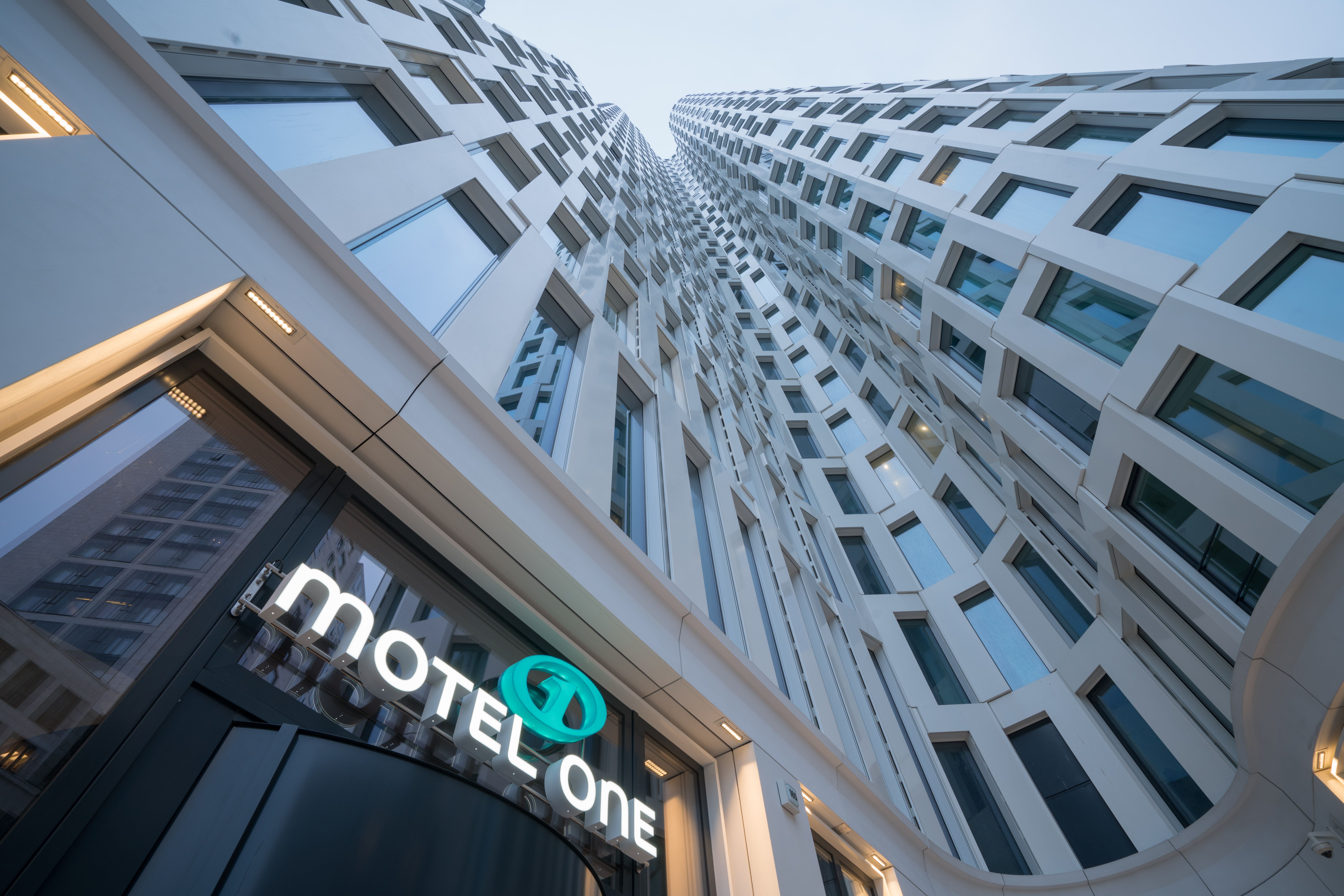 Blick von unten auf die moderne, hohe Fassade eines Hotels mit vielen Fenstern und dem leuchtenden Schriftzug „Motel One“ am Eingang.