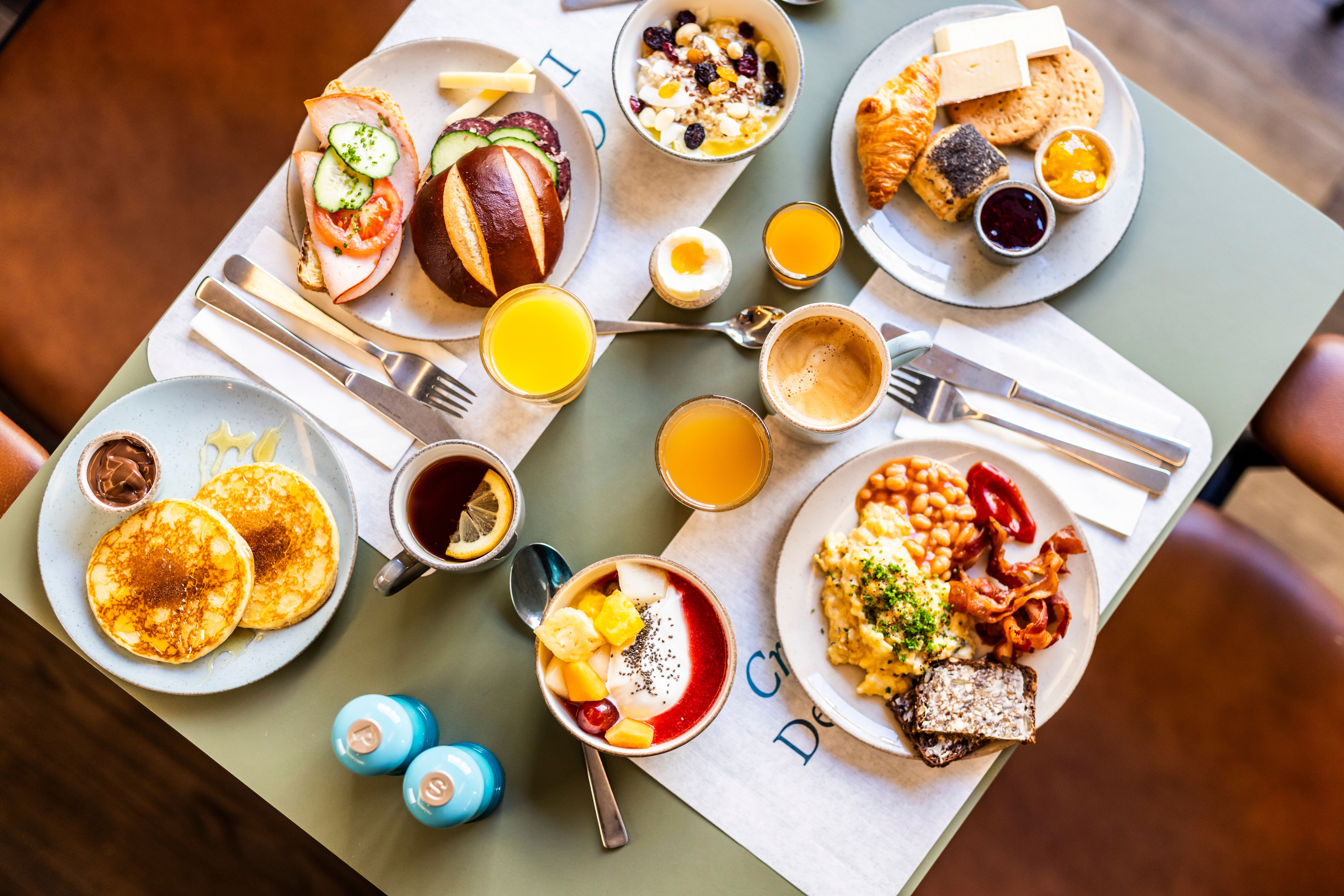 Vielseitiges Frühstück mit Pancakes, Rührei, Bacon, Brötchen, Croissant, Joghurt mit Früchten, frischem Obst, Marmelade, Säften und Kaffee, liebevoll angerichtet auf einem Tisch.