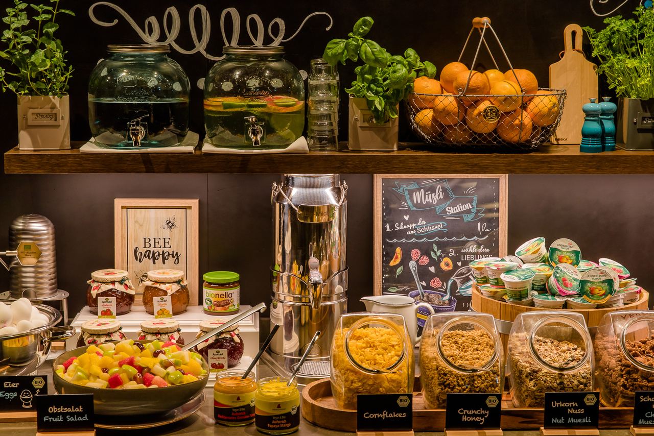 Vielseitiges Frühstücksbuffet mit frischem Obstsalat, verschiedenen Müslisorten, Honig, Marmelade, frischen Orangen, Kräutern und Getränkespendern für Wasser und infused Water. Gemütliche und einladende Atmosphäre.