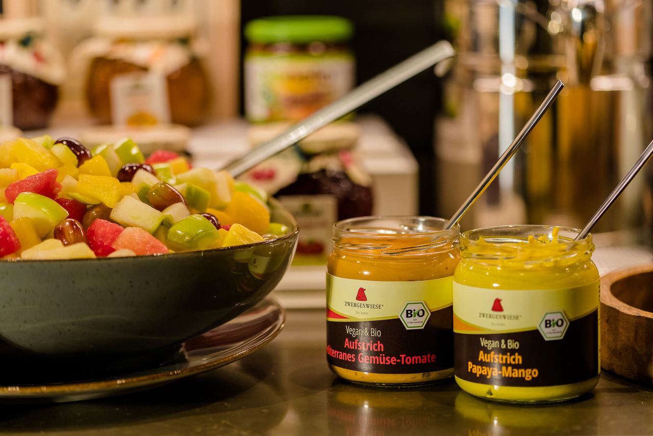 Schale mit frischem gemischtem Obstsalat und zwei Gläsern mit veganen, biologischen Aufstrichen in den Geschmacksrichtungen Gemüse-Tomate und Papaya-Mango.