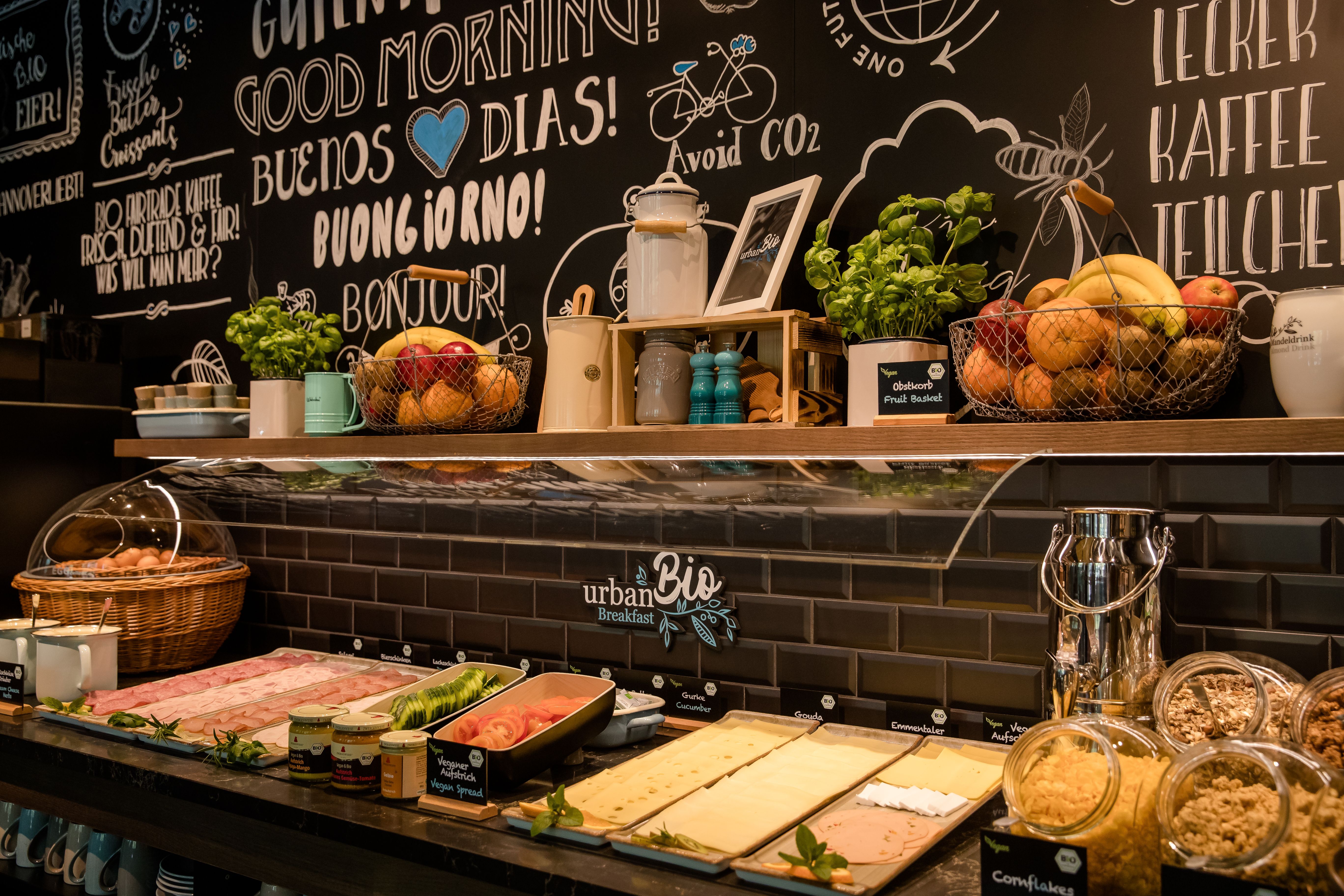 Frühstücksbuffet mit frischem Obst, Aufschnitt, Käse, Müsli und Kräutern vor einer dekorativen Wand mit internationalen Begrüßungen und nachhaltigen Botschaften.