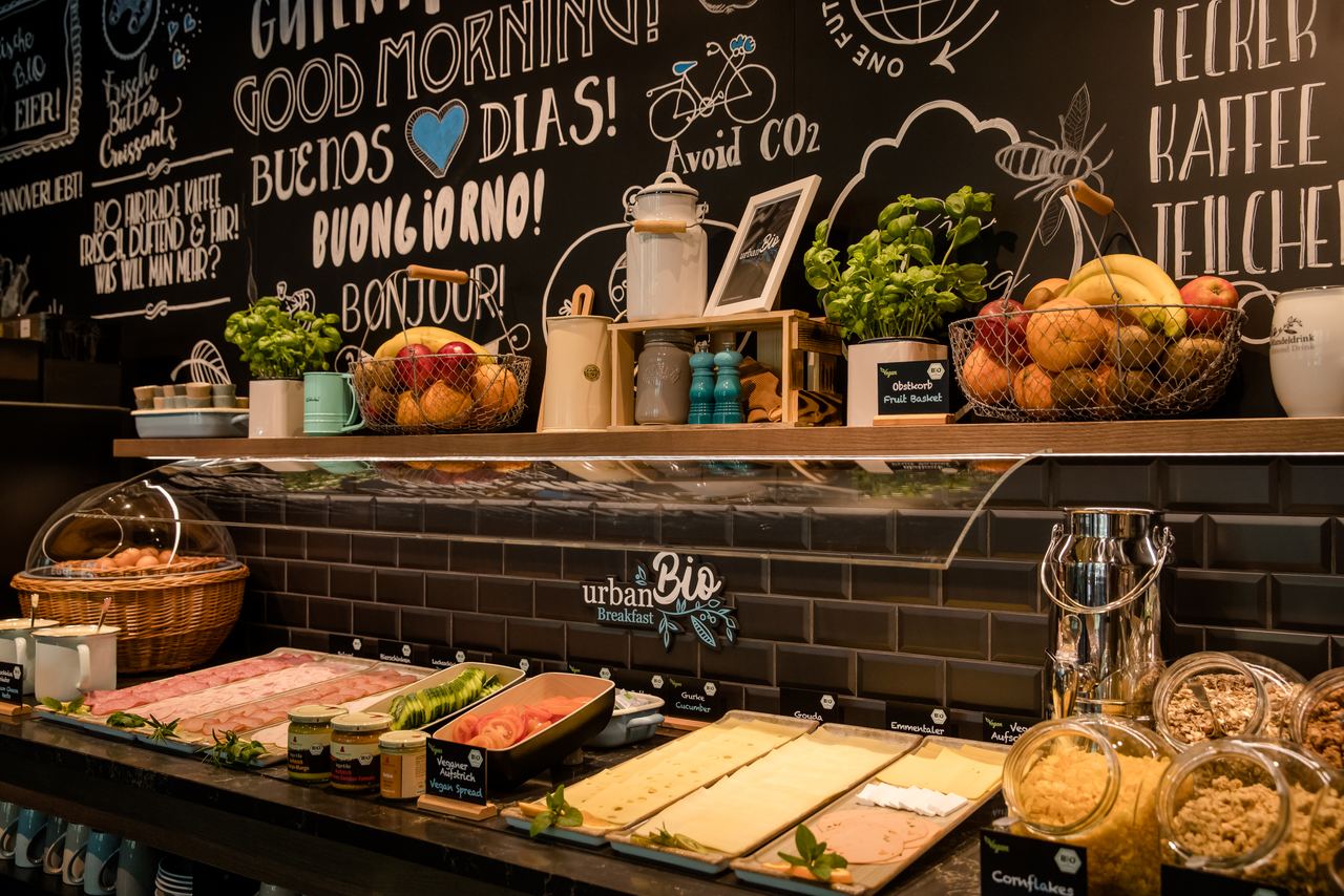 Frühstücksbuffet mit frischem Obst, Aufschnitt, Käse, Müsli und Kräutern vor einer dekorativen Wand mit internationalen Begrüßungen und nachhaltigen Botschaften.